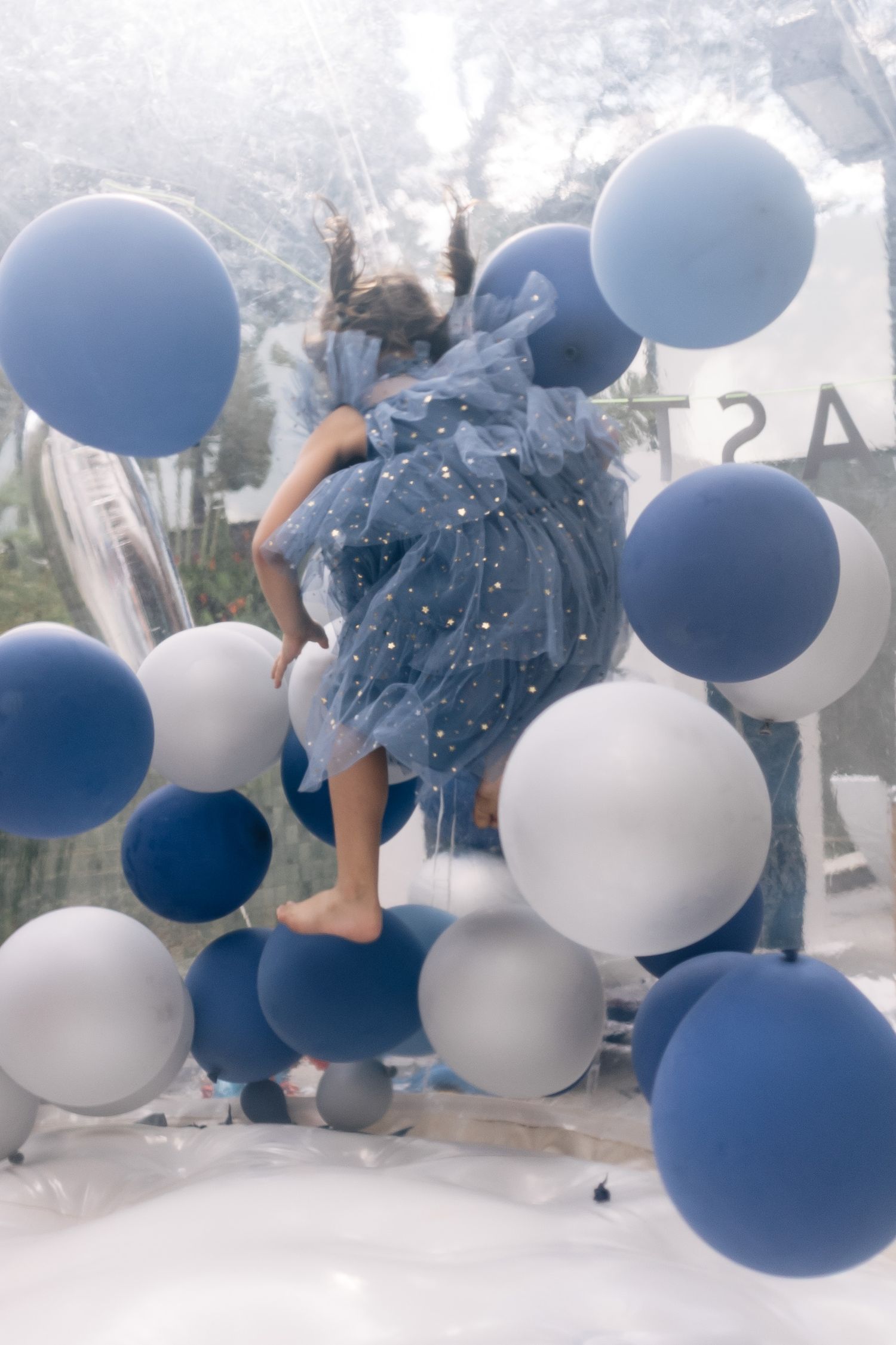 Girl in blue tulle dress jumping among blue, white, and gray balloons in a dreamy, ethereal setting.