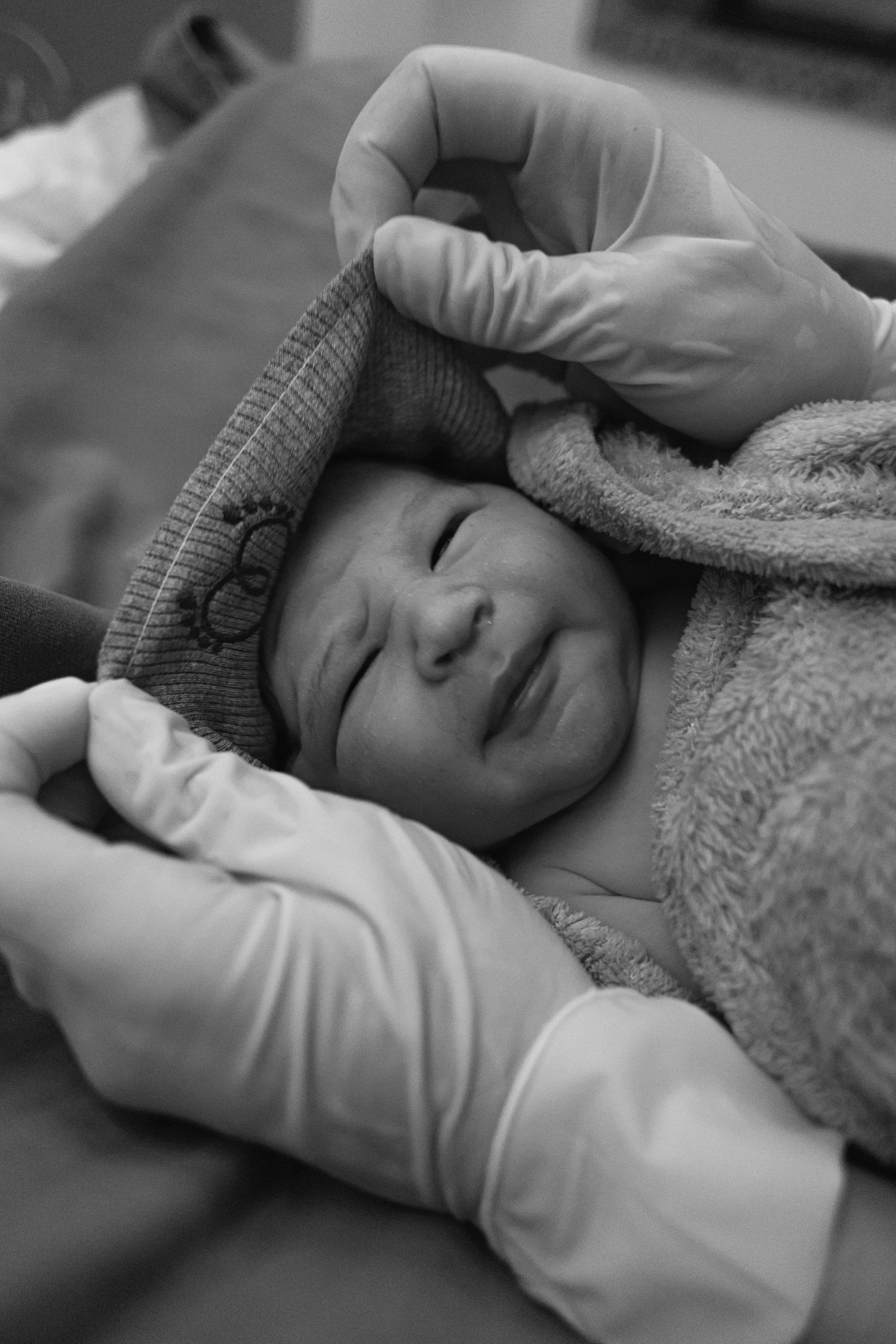Black and white photo of a newborn wearing a knit cap while being examined by gloved hands.