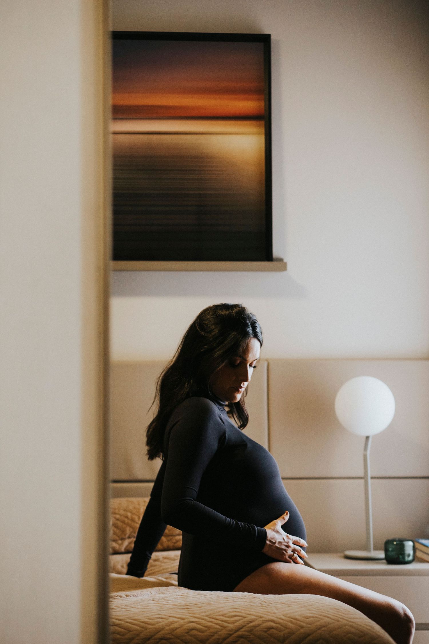 Artistic maternity photo of pregnant woman sitting on bed beneath sunset artwork.