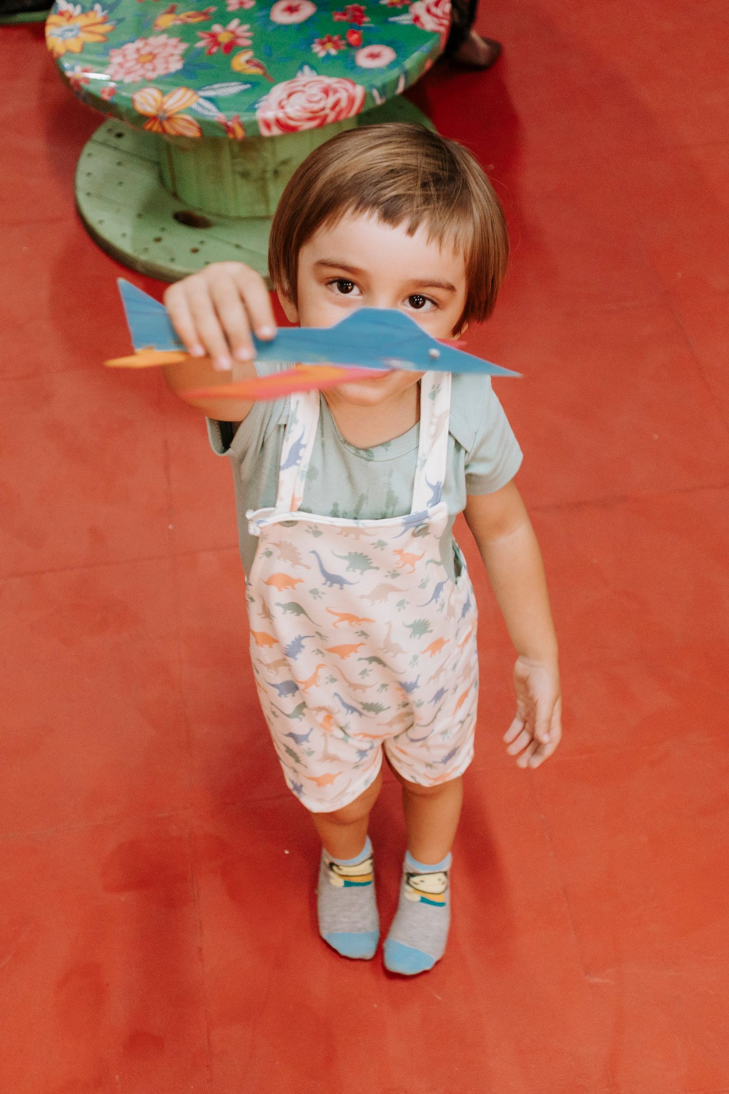 A toddler in overalls holds a blue toy fish while standing on a red floor.