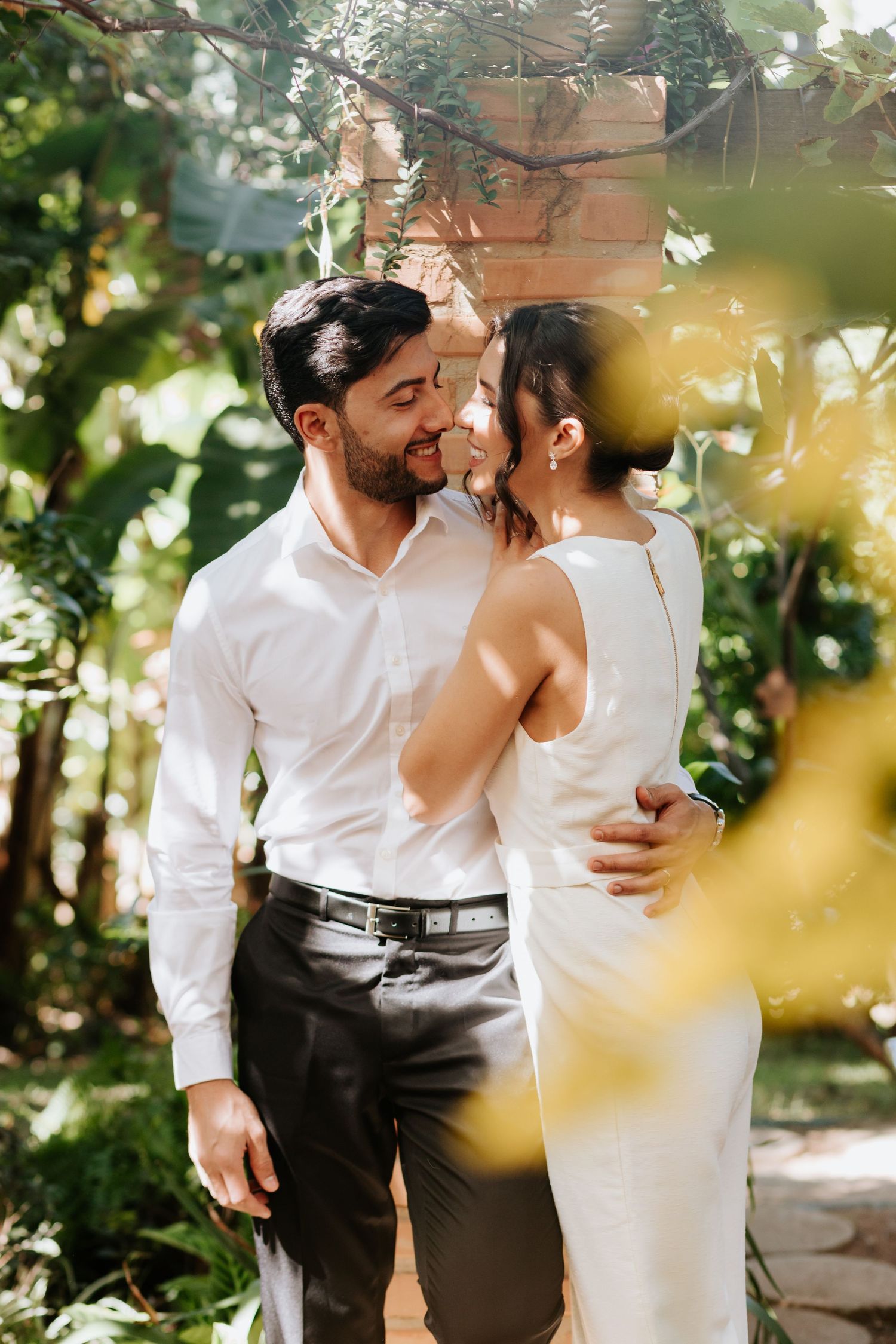 A couple in white clothing embraces in a sun-dappled garden setting during an intimate photoshoot.