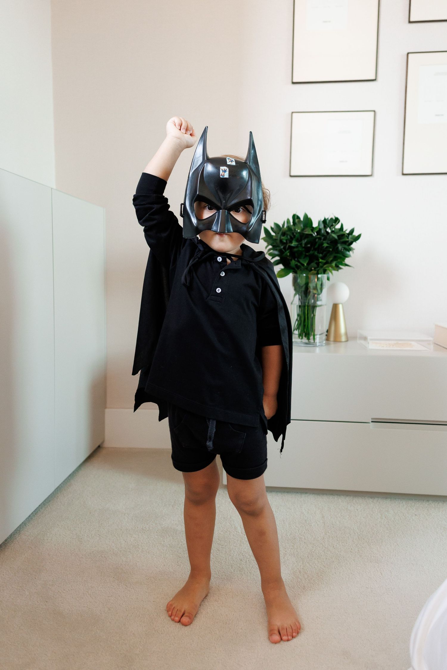 Child dressed in Batman costume with mask and cape, striking a superhero pose with fist raised indoors.