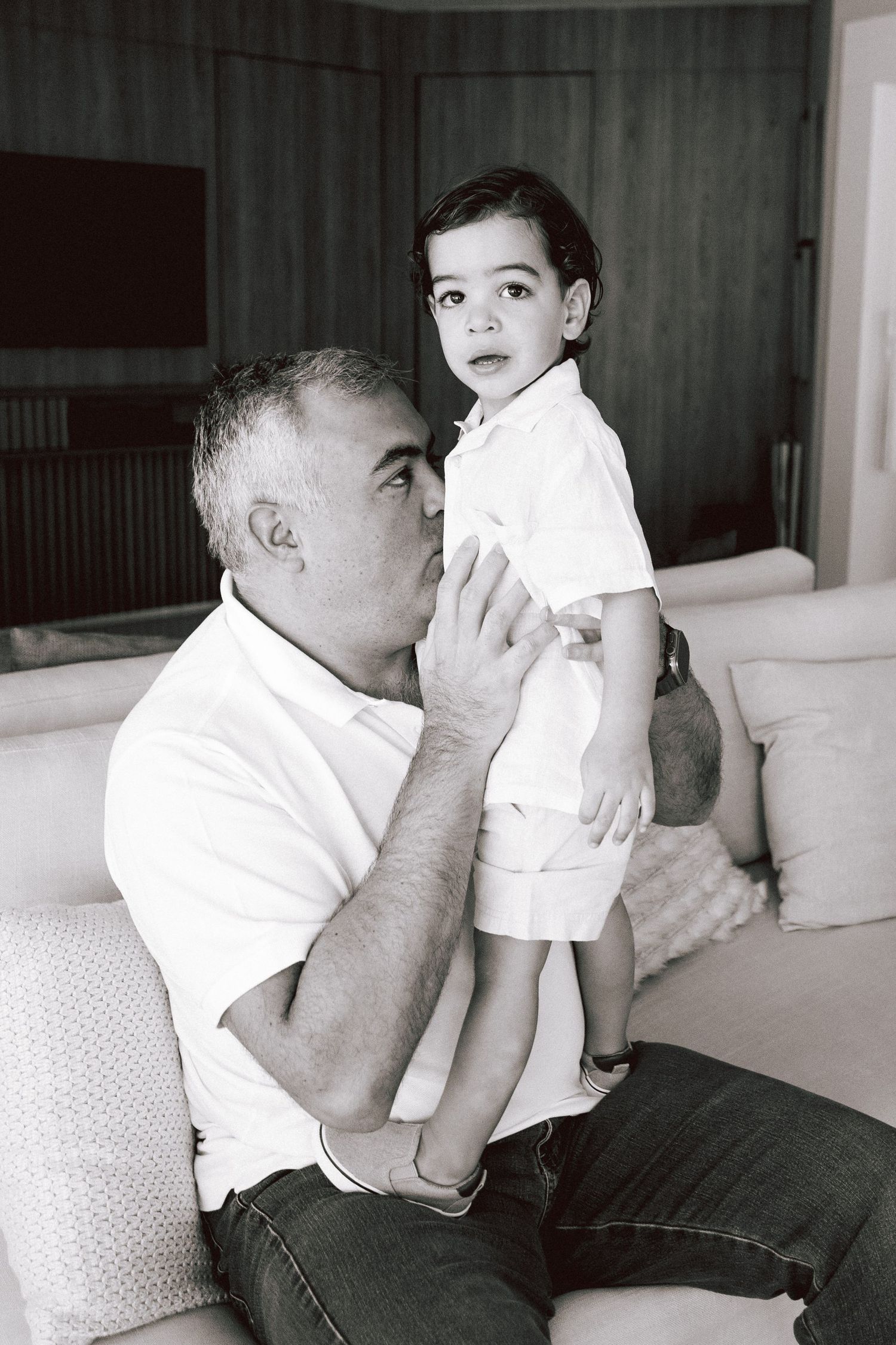 Black and white photo of a man kissing a toddler boy while holding him on a couch indoors.