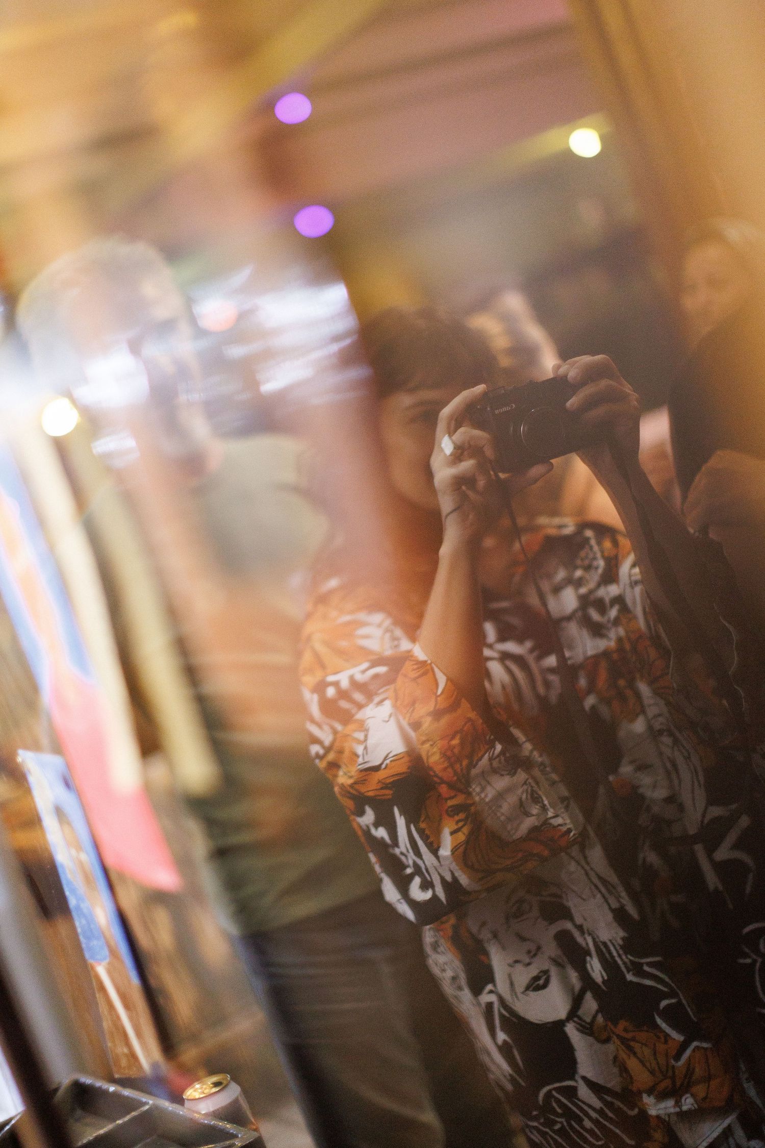 Person in graphic shirt photographing their reflection in a store window at night with colorful bokeh lights in background.