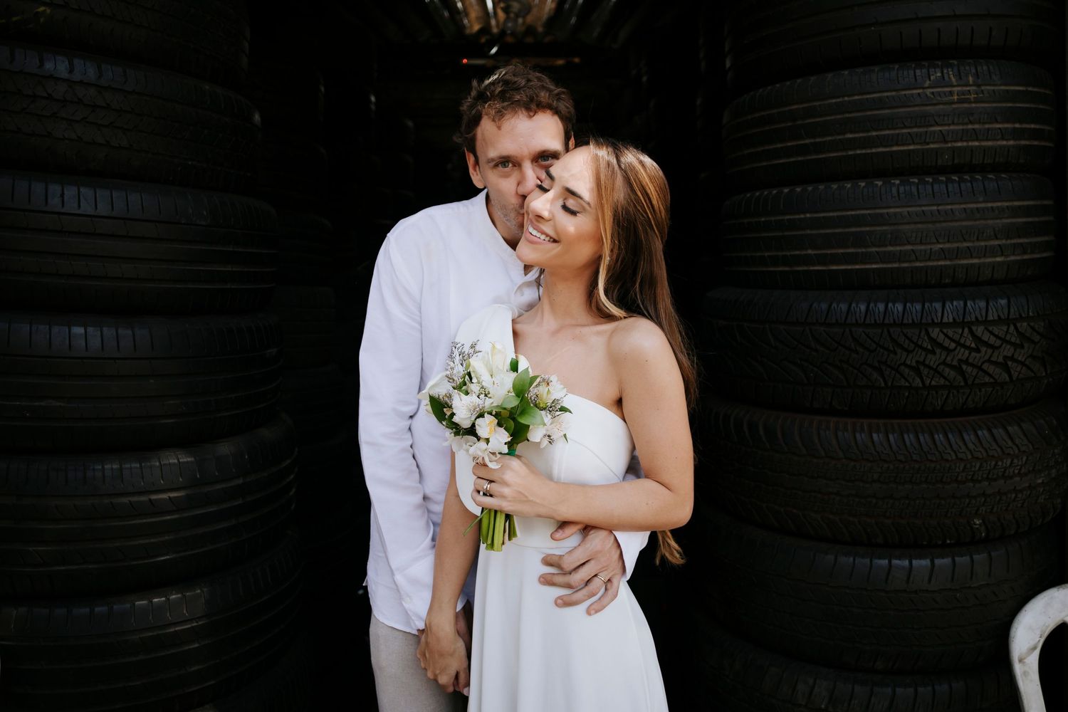 A couple in white wedding attire sharing a romantic moment against a backdrop of stacked tires.