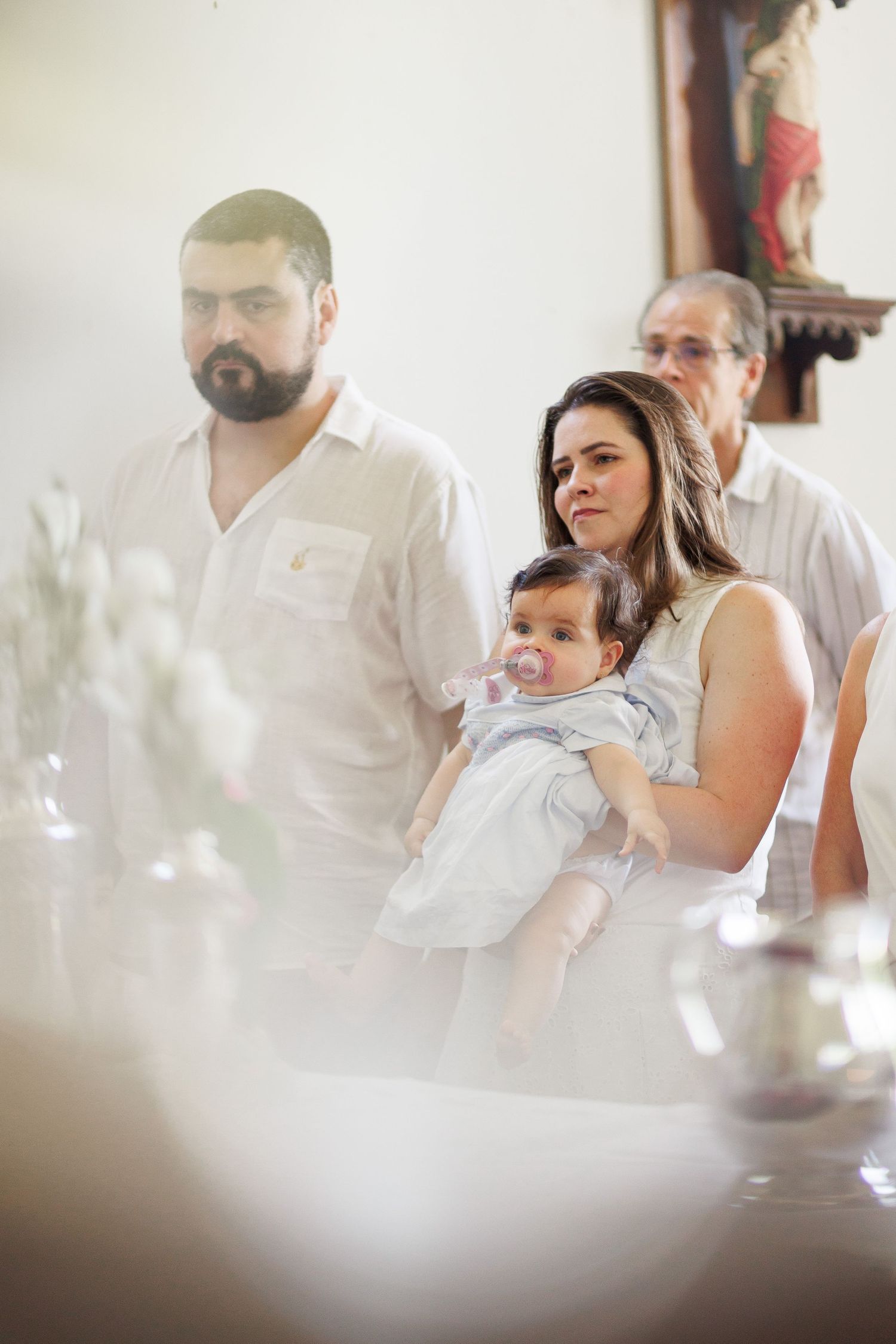 Family at a baptism ceremony, mother holding baby with pacifier, father and grandfather standing nearby in a church.