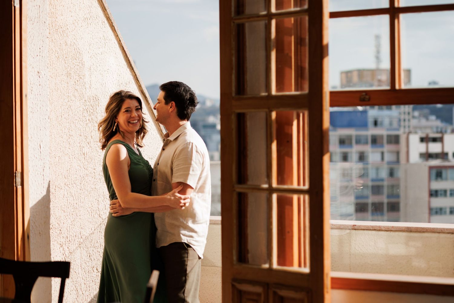 A couple shares an intimate moment in a sunlit room with wooden window frames and urban views.