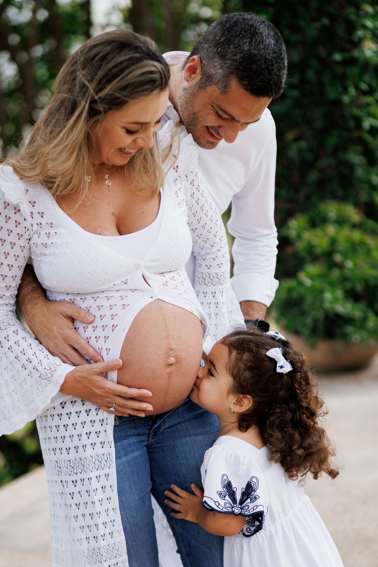 A family shares tender moments during a maternity photoshoot outdoors, wearing coordinated white outfits.