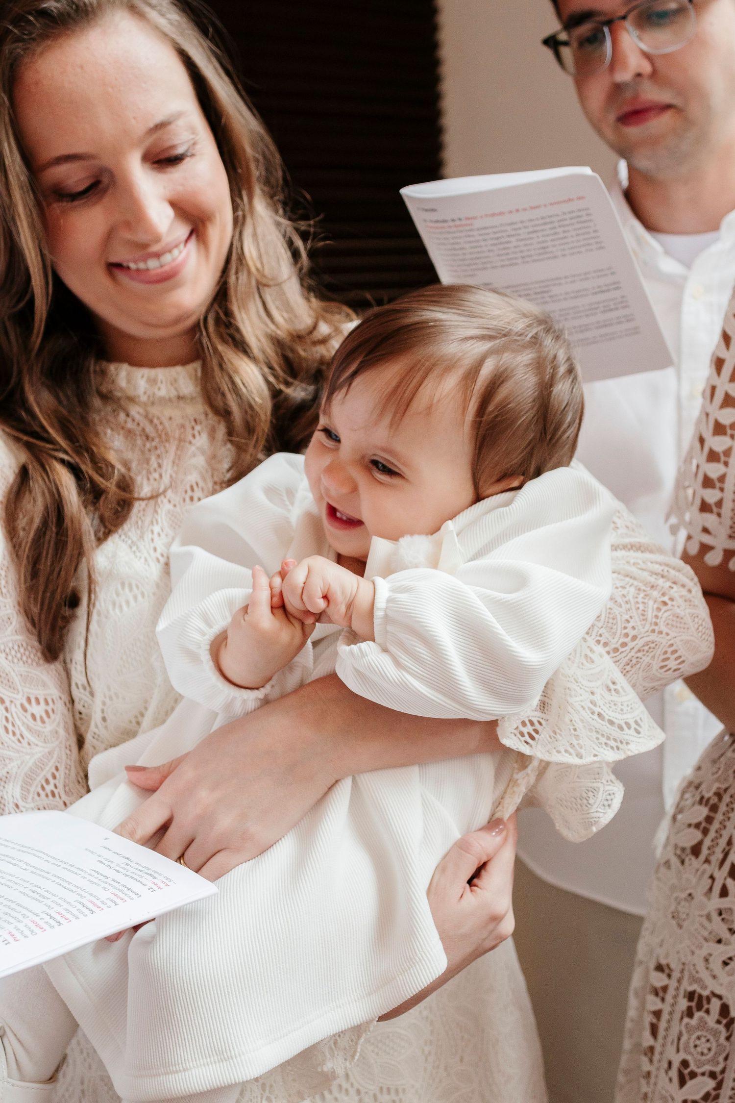 A baby in a white christening gown smiles while being held during a religious ceremony.