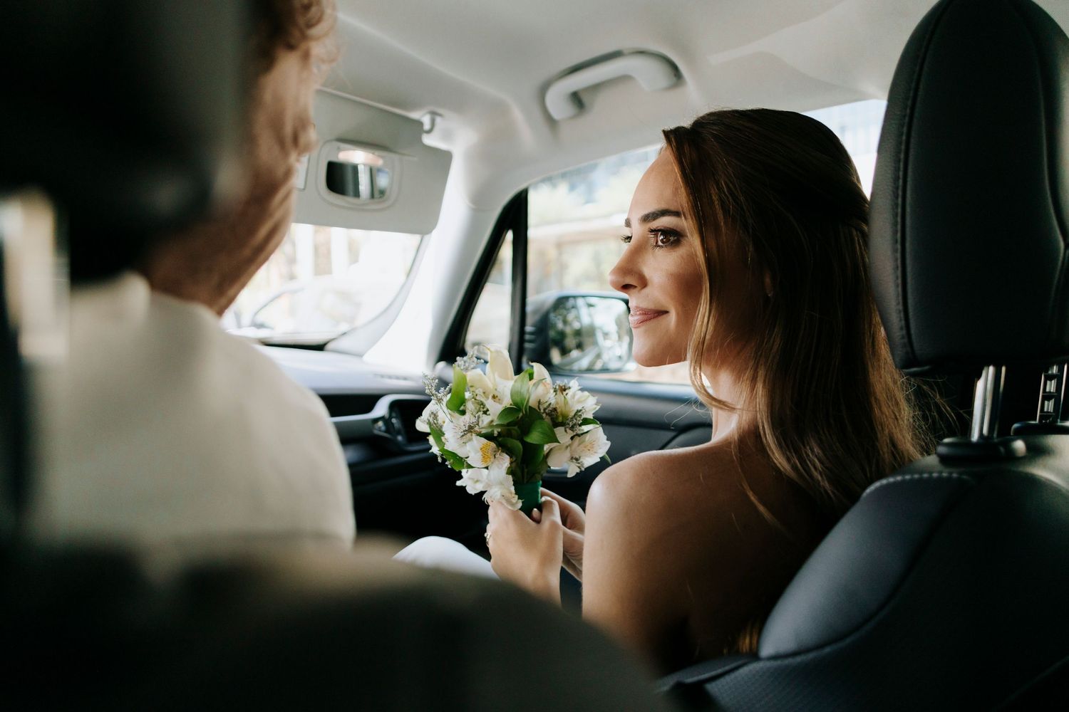 Bride with bouquet smiling at partner while sitting in car on wedding day.