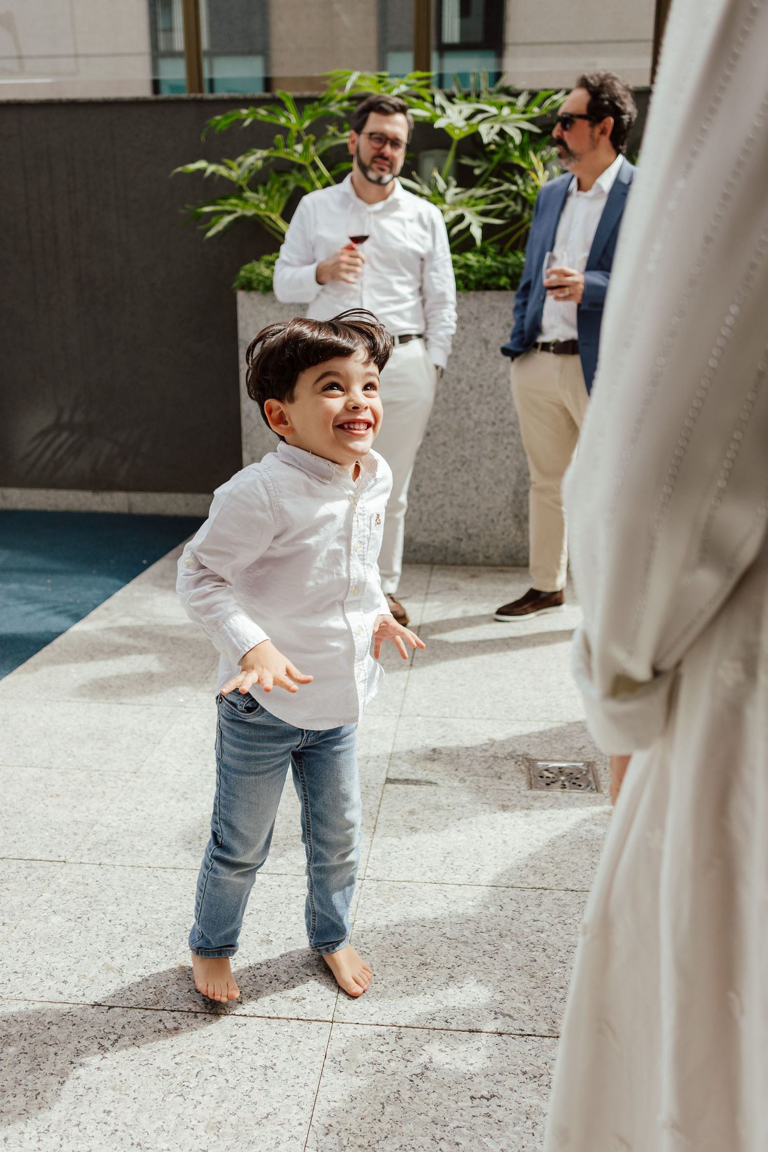 A young child in a white shirt and jeans plays joyfully between white pillars in an outdoor courtyard.
