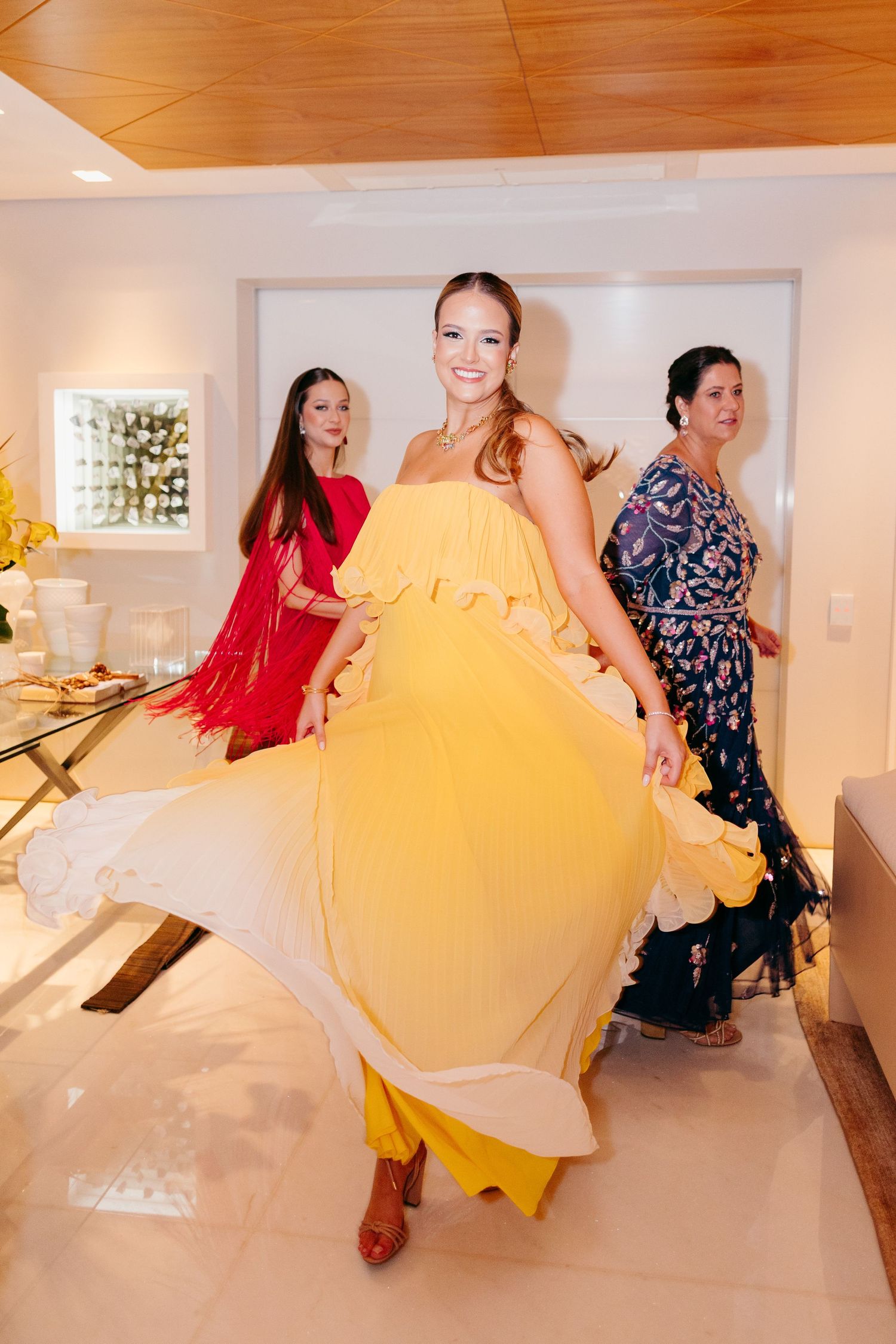 Three women in colorful dresses twirl and dance in an elegant indoor setting with wooden ceiling and decorative lighting.