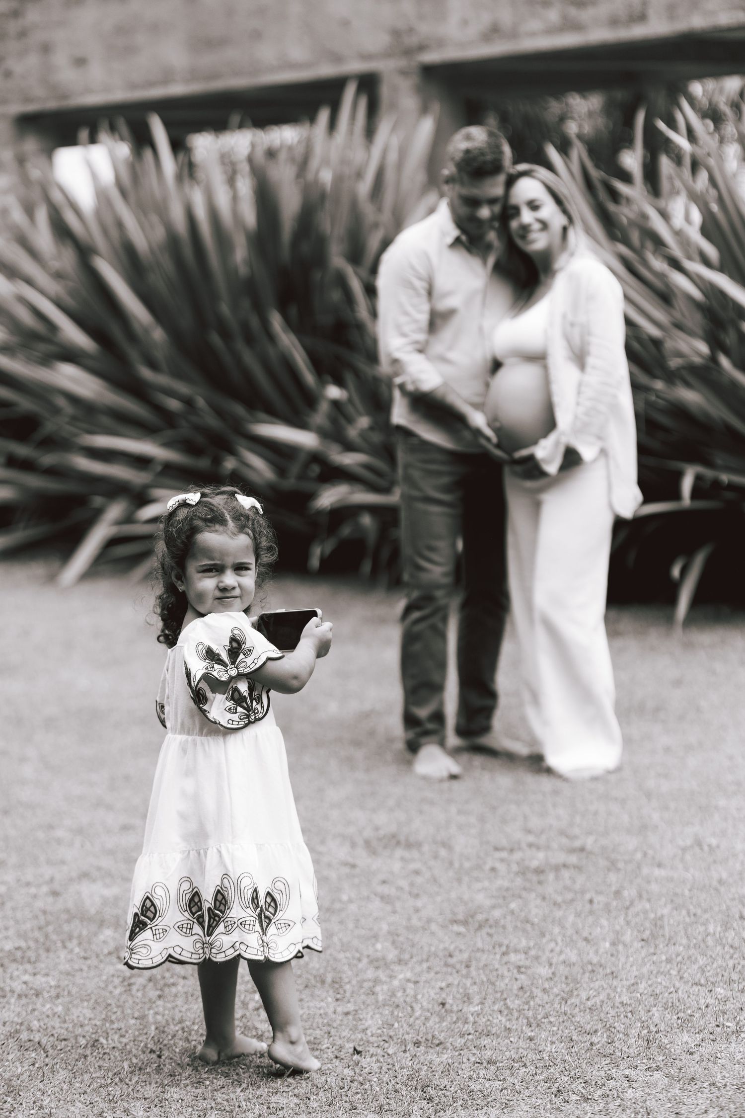 A young child in a white embroidered dress plays and twirls outdoors on gravel near some plants.