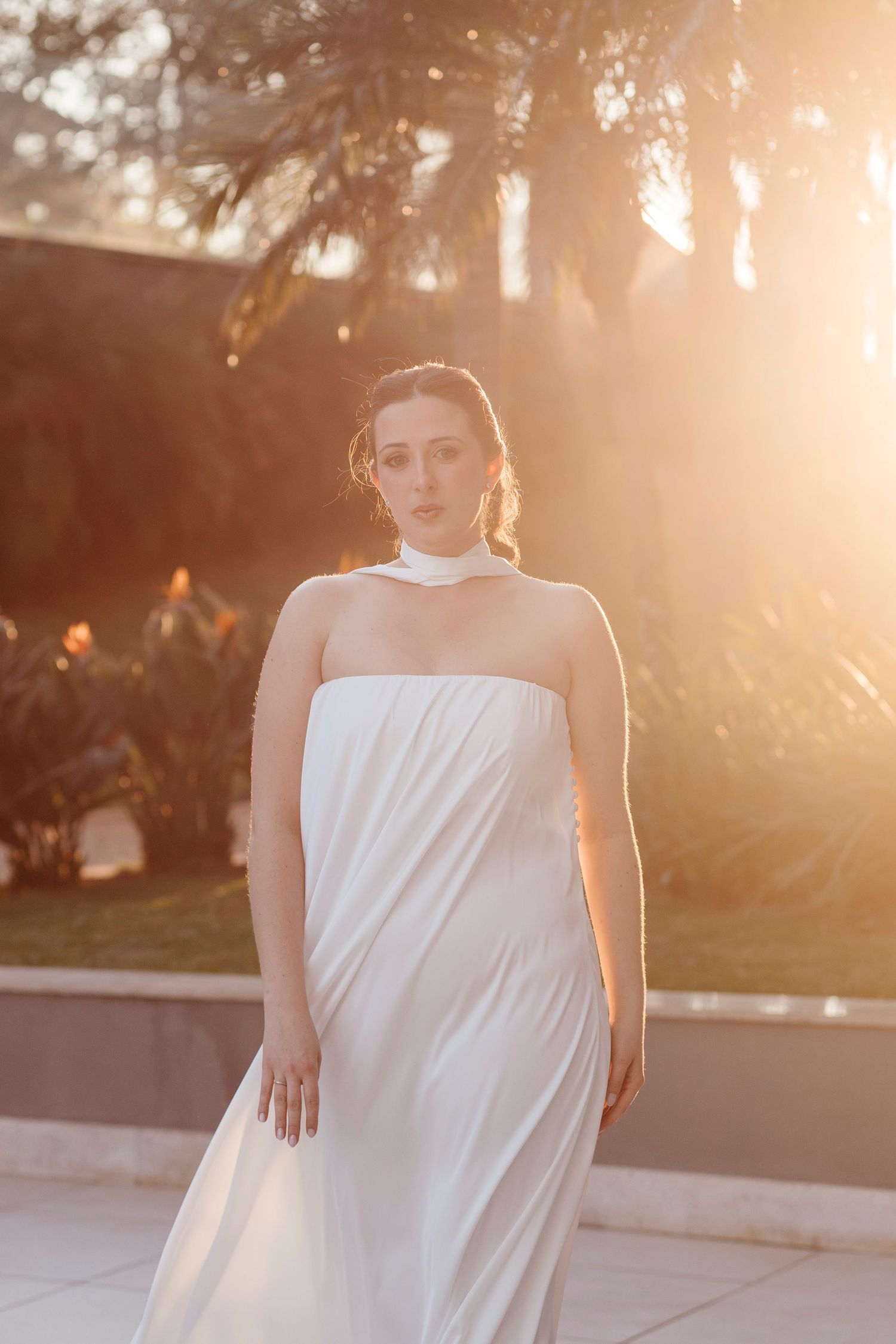 A figure in a flowing white dress poses outdoors during golden hour sunset creating a dreamy silhouette effect.