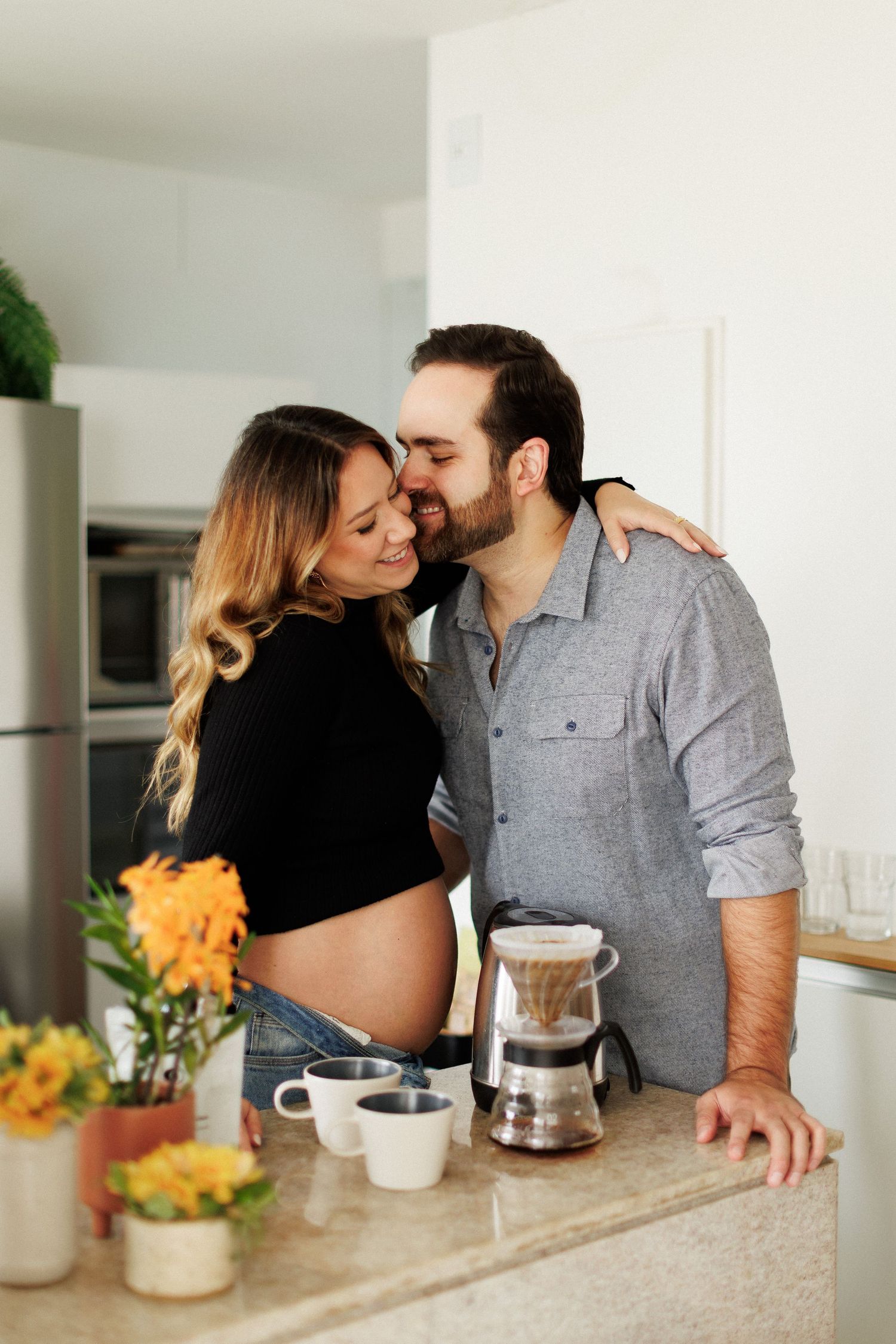 Happy couple sharing a tender kiss in kitchen, pregnant woman with exposed baby bump standing by coffee maker.