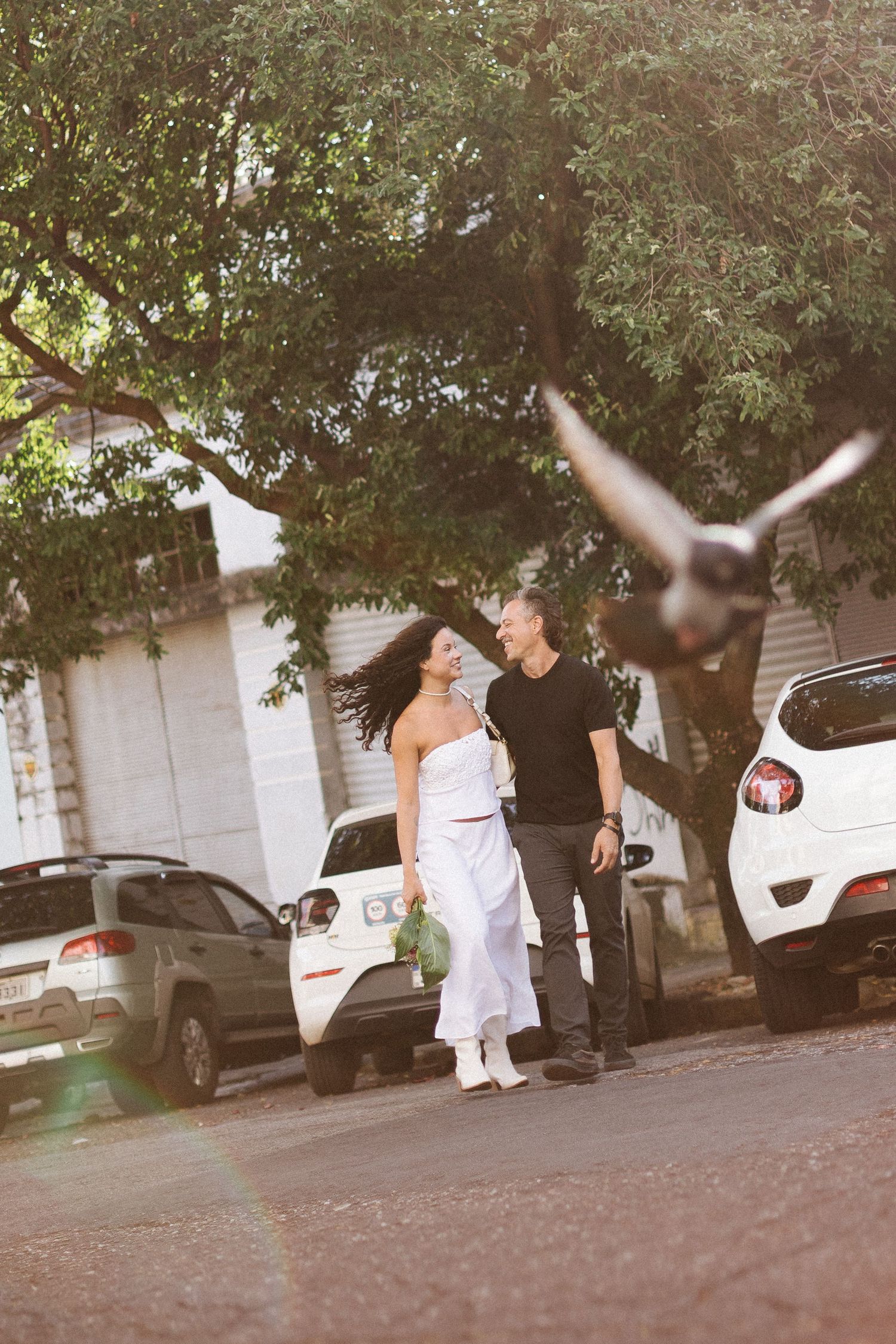 Couple walking and laughing on a sunny street with parked cars and trees, as a pigeon flies past in the foreground.