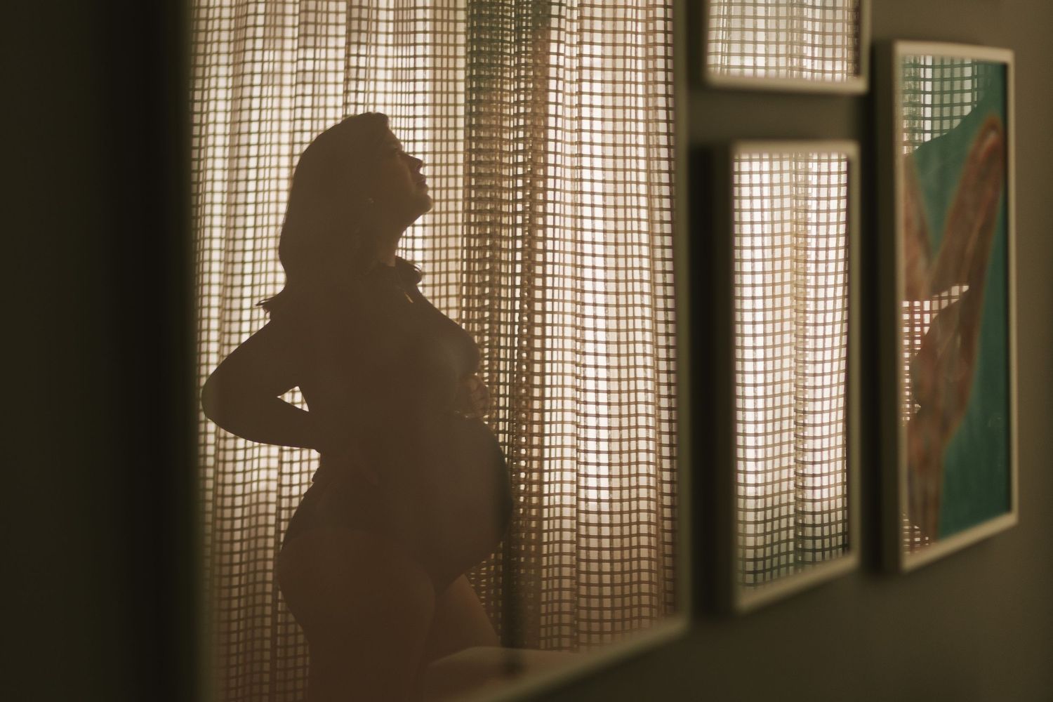 Silhouette of a pregnant woman standing by sheer curtains with warm backlight, framed art visible on wall.