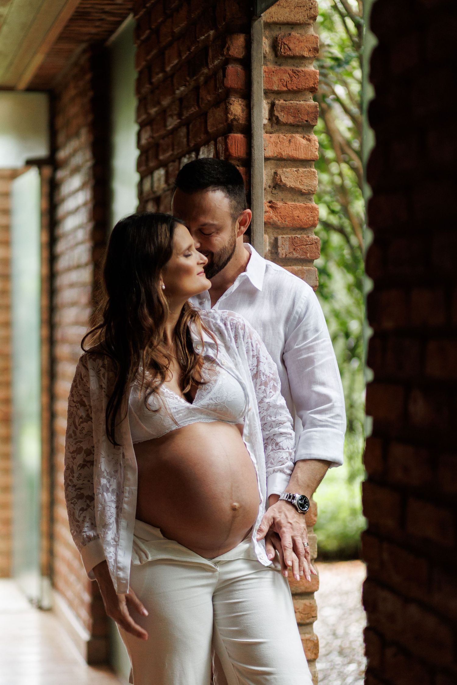 Couple embracing outdoors, pregnant woman in white lace top with partner in white shirt against brick wall.