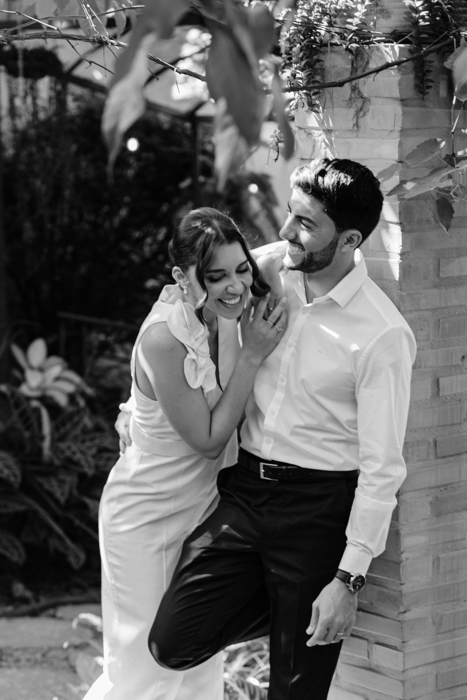 Black and white photo of a smiling bride and groom leaning against a brick wall surrounded by lush greenery.