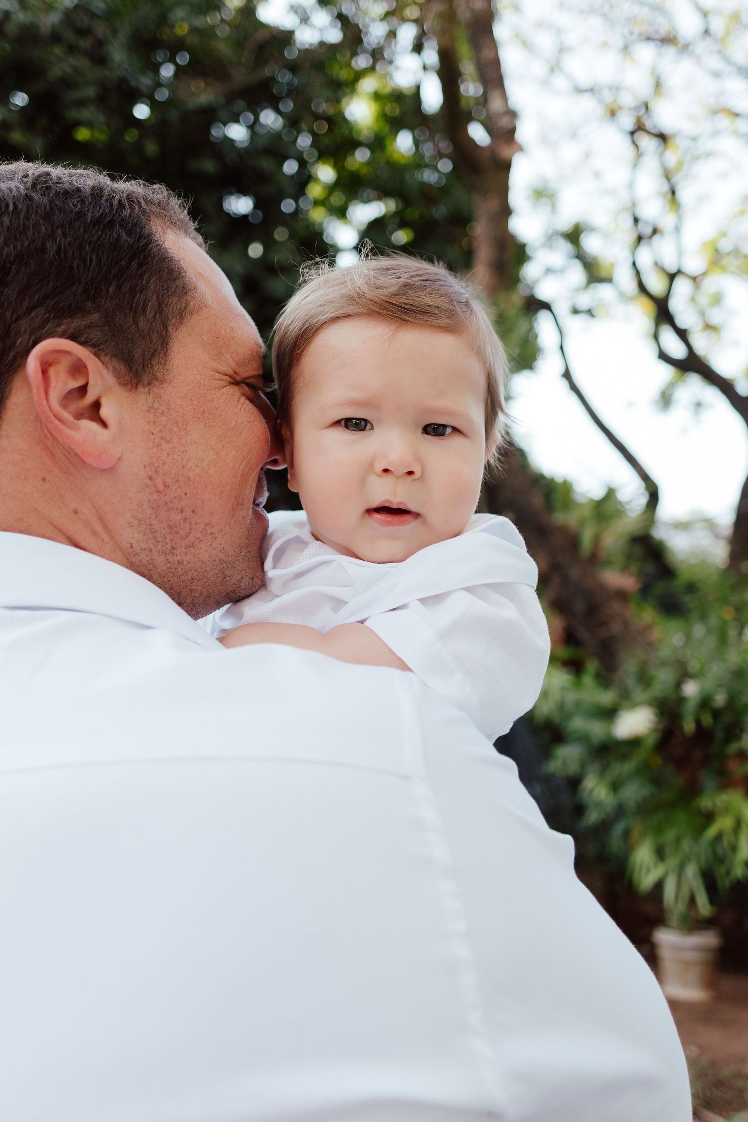 A tender moment outdoors as a parent in white holds a small child against lush greenery in the background.