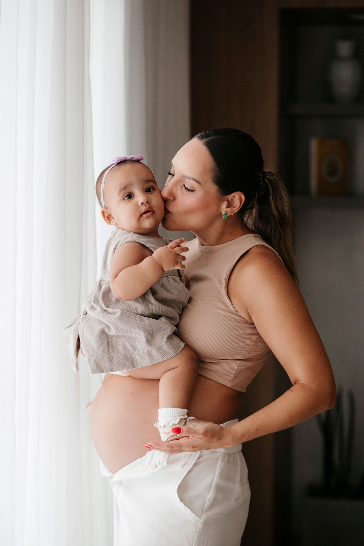 A pregnant woman in neutral tones kisses a baby while standing by a bright window.