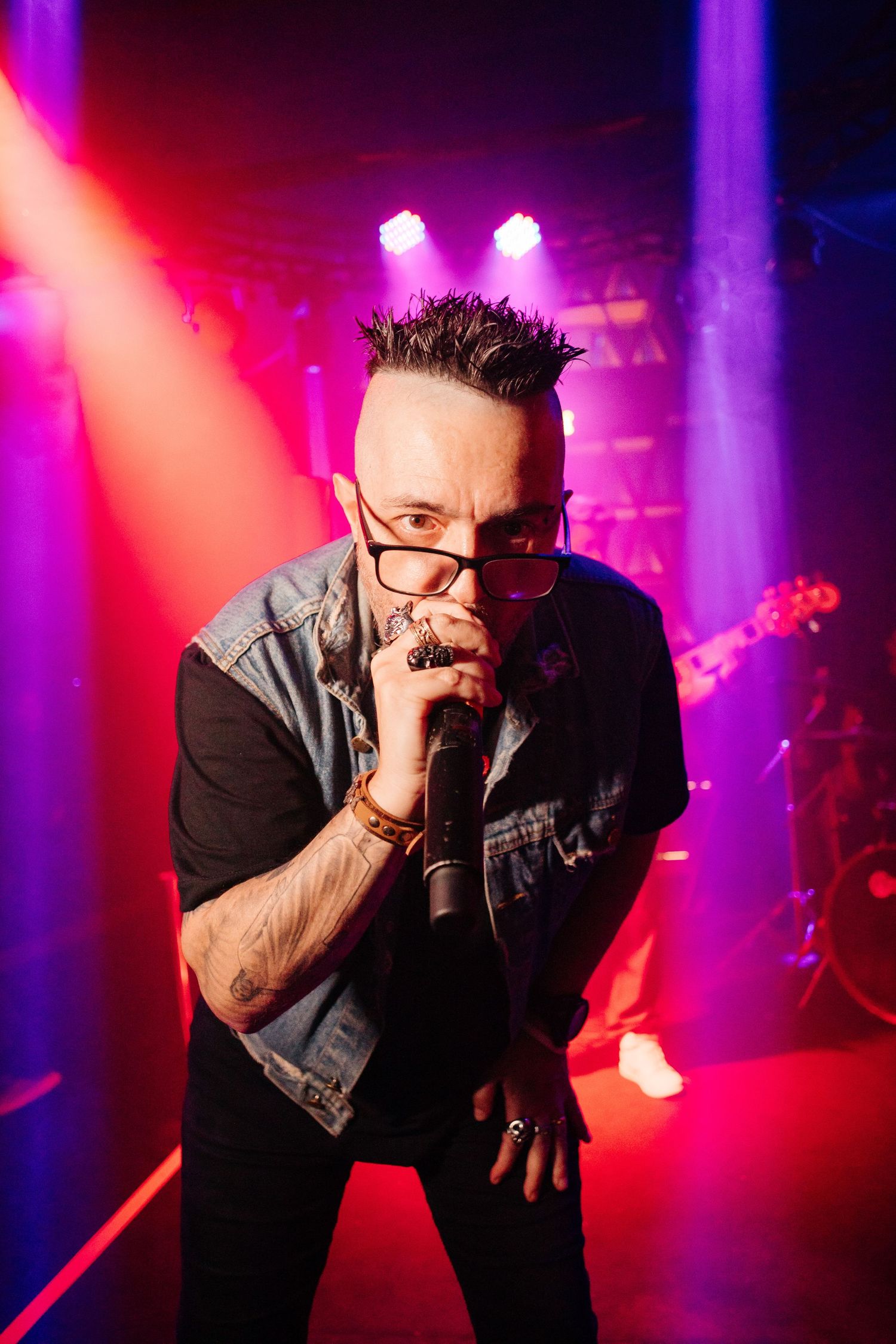 Musician performs passionately with harmonica in dark concert venue illuminated by dramatic red and purple stage lighting.
