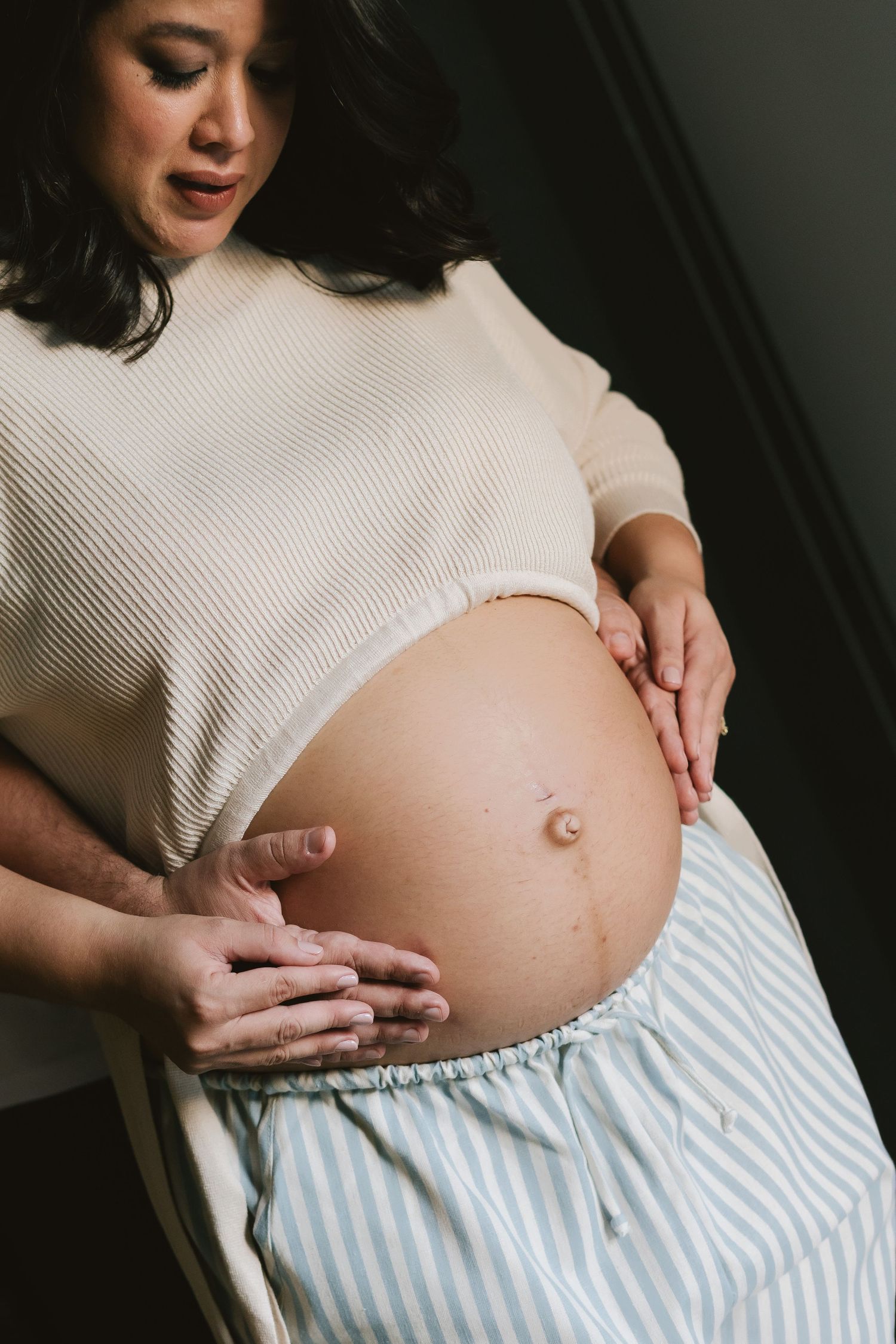 Pregnant woman in white top and striped skirt cradling her bare baby bump with a partner's hands visible.