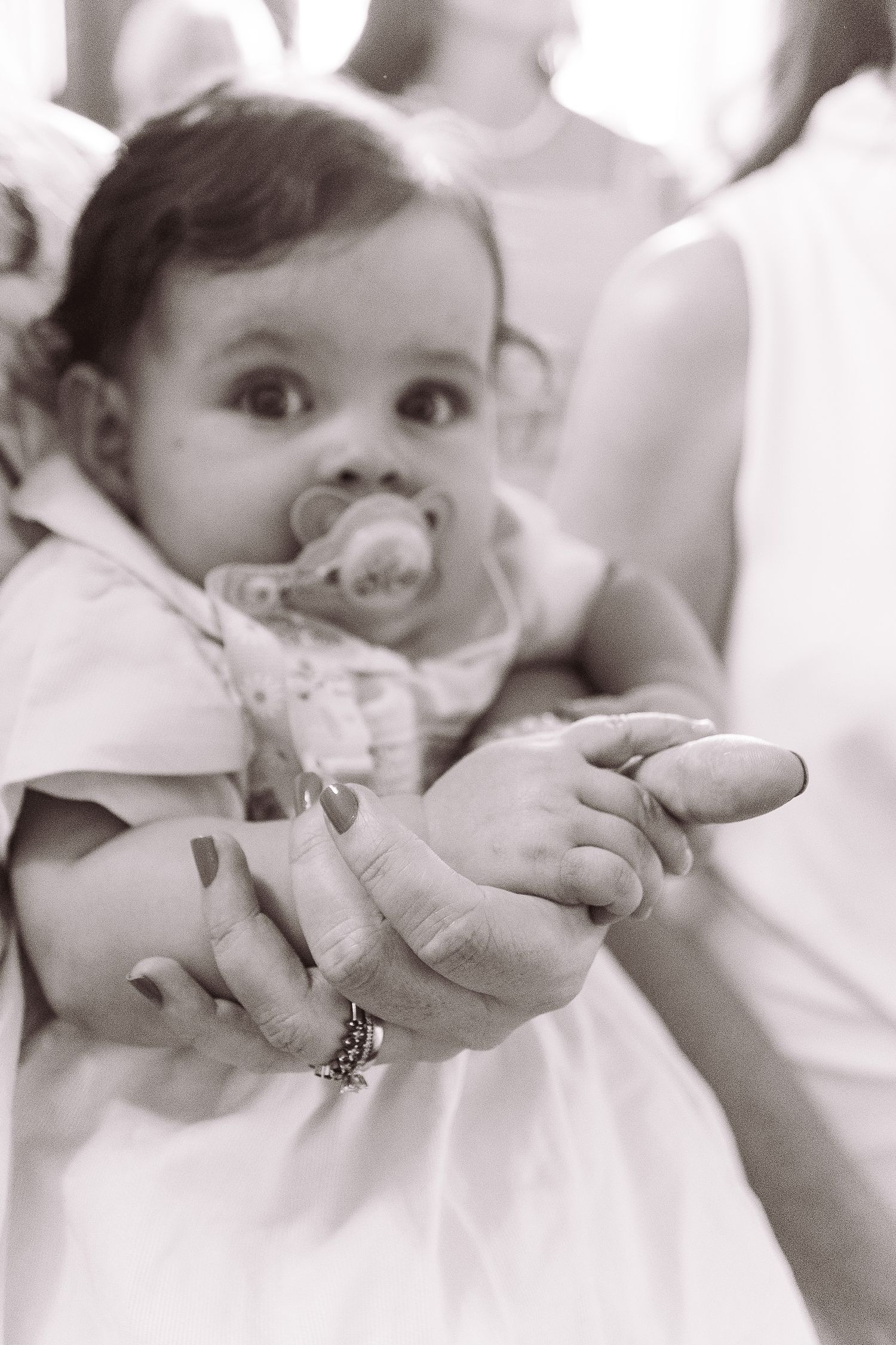 Black and white photo of a baby with a pacifier being held by an adult woman wearing a bracelet and nail polish.