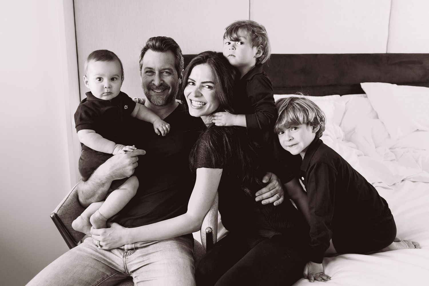 Black and white family portrait of parents with three young children sitting together on a bed, smiling warmly.