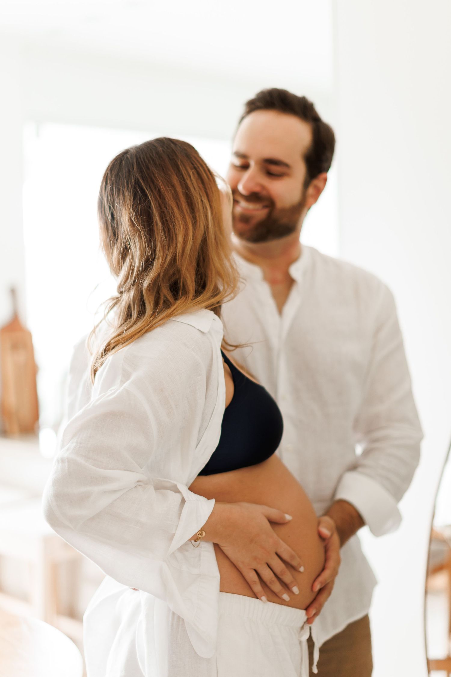 A smiling bearded man in a white shirt tenderly holds the baby bump of a pregnant woman in a bright, airy room.