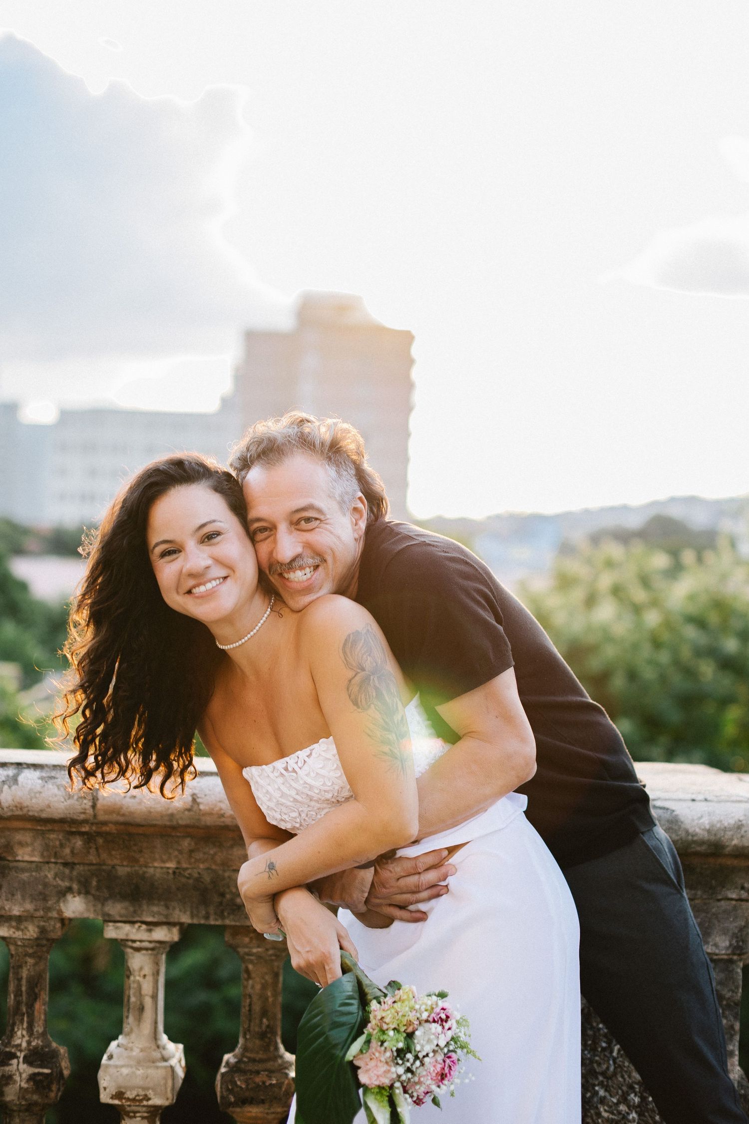Smiling bride in white dress holds bouquet as groom hugs her from behind on a sunlit balcony.
