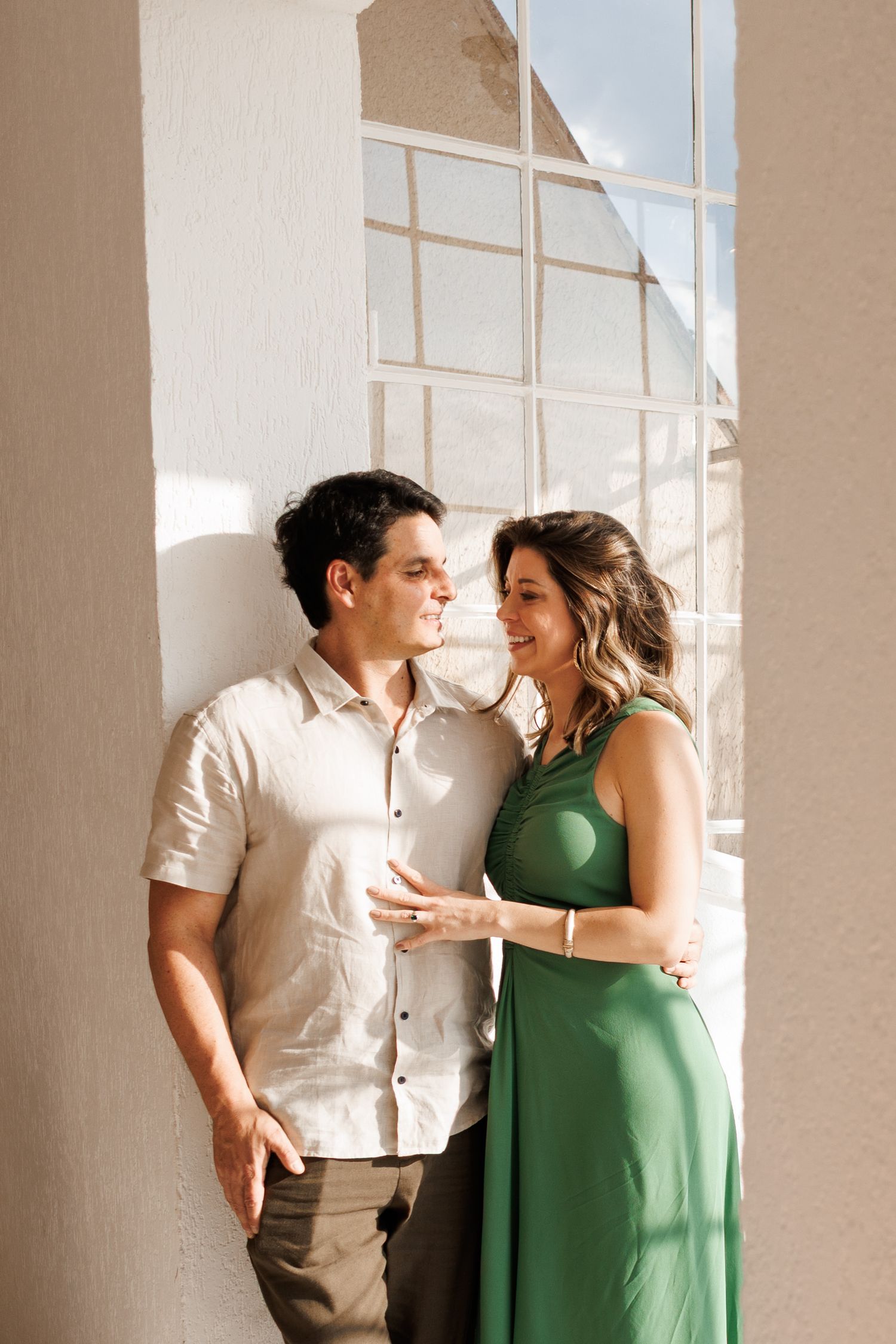 A couple in white and green outfits share intimate moments against a bright sunlit wall with windows.
