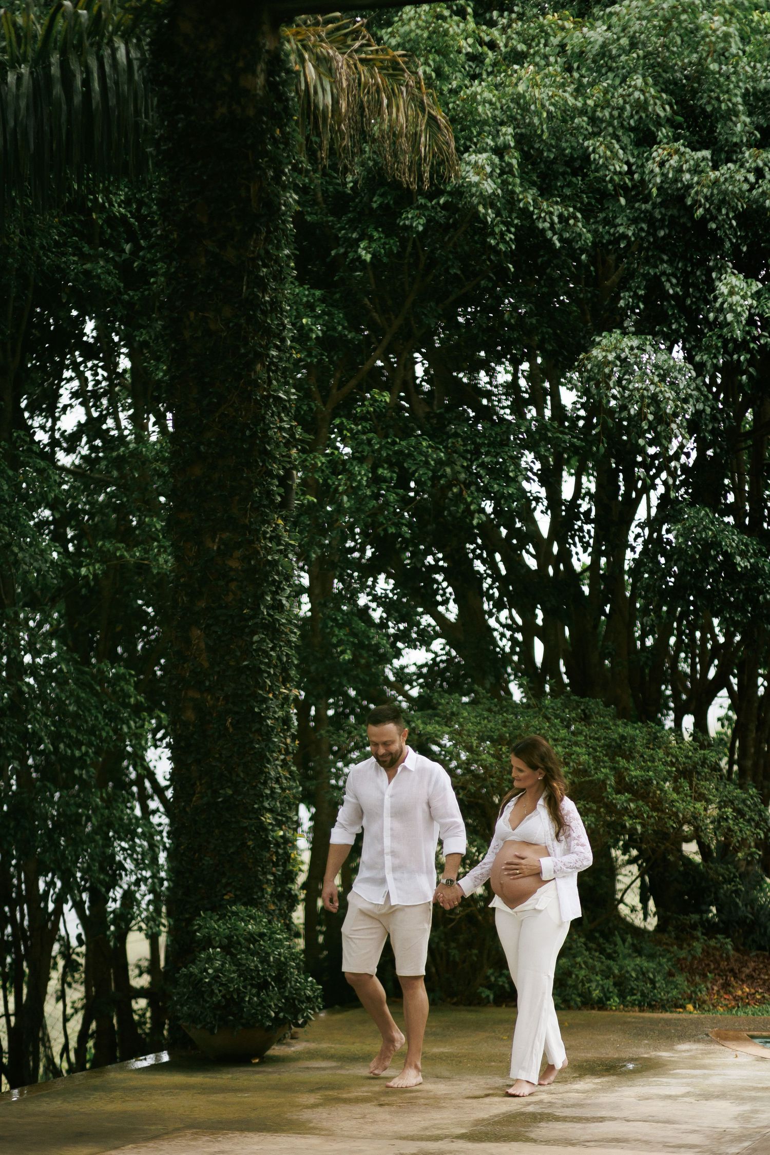 Pregnant couple in white outfits holding hands while walking through a lush green garden setting.