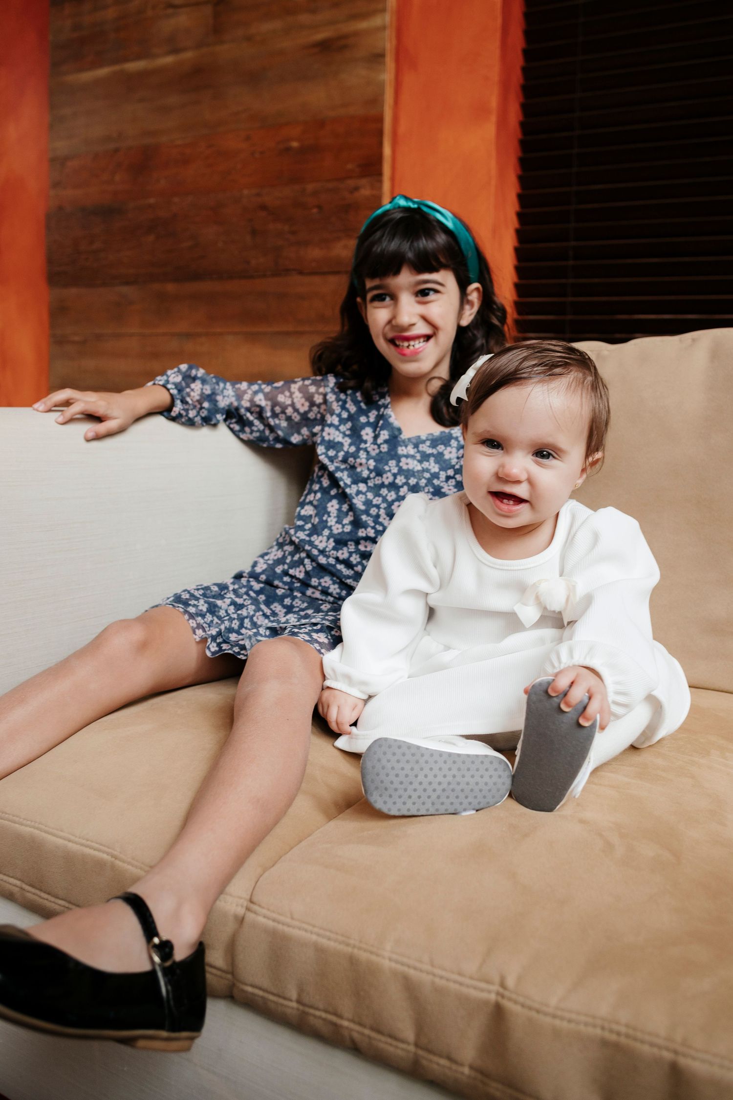Children relaxing on tan couch with girl in floral dress and baby in white outfit.