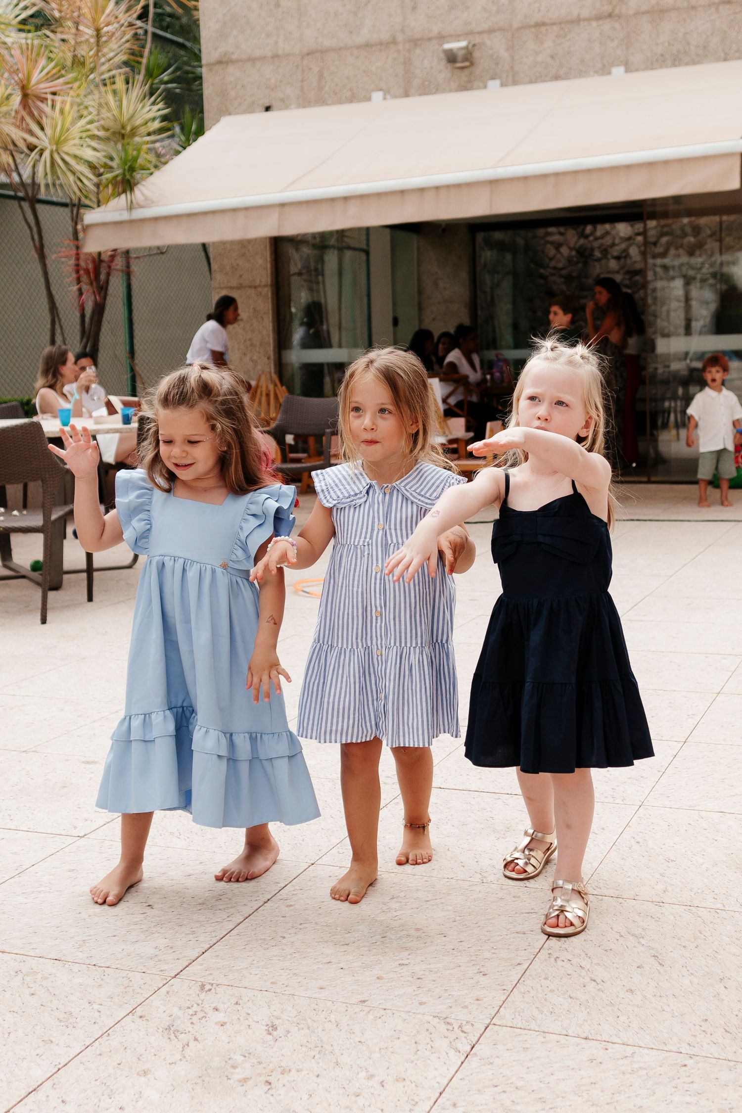 Three young children in summer dresses dancing together on an outdoor patio.