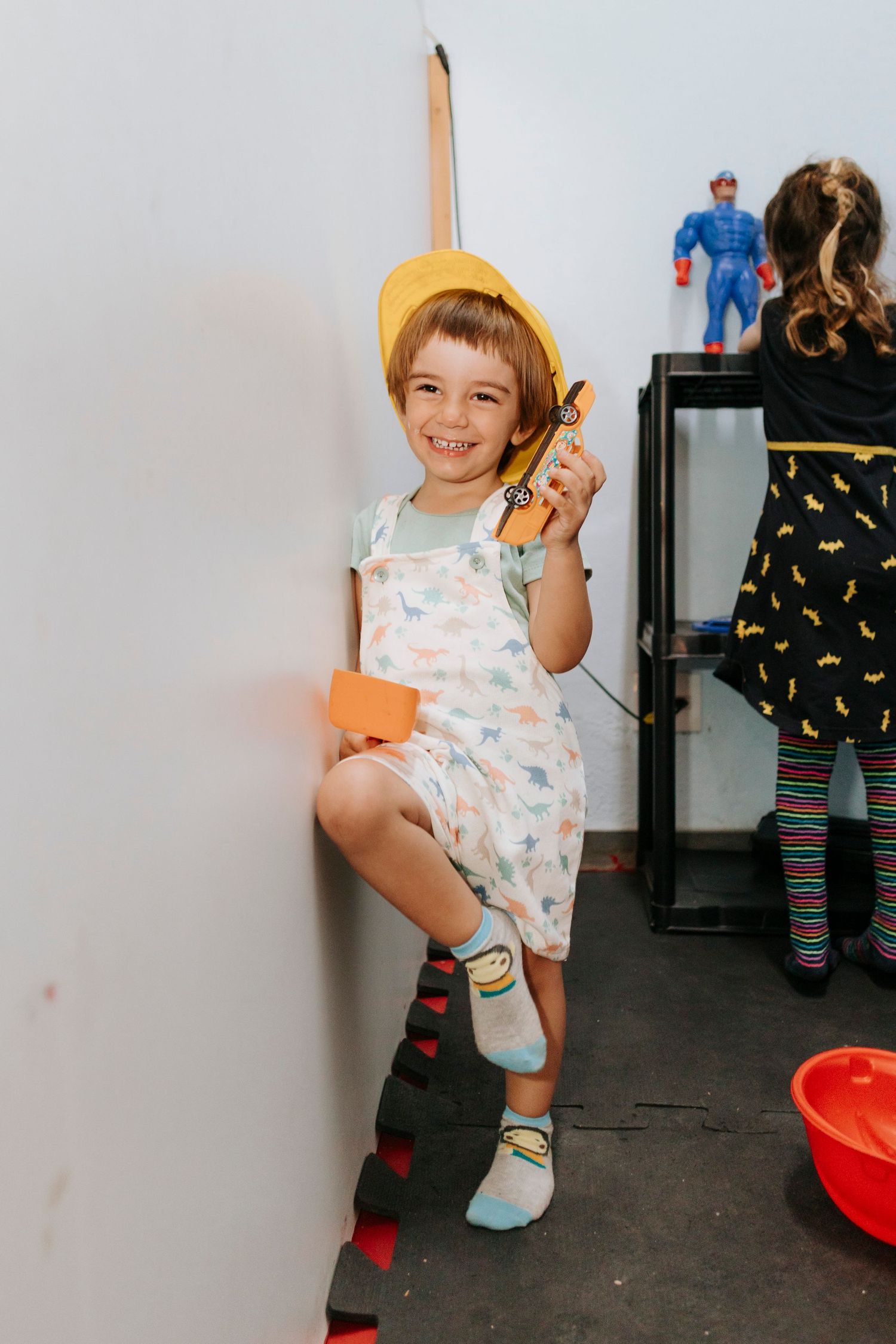 A child wearing a yellow hat and patterned overalls poses playfully against a white wall.