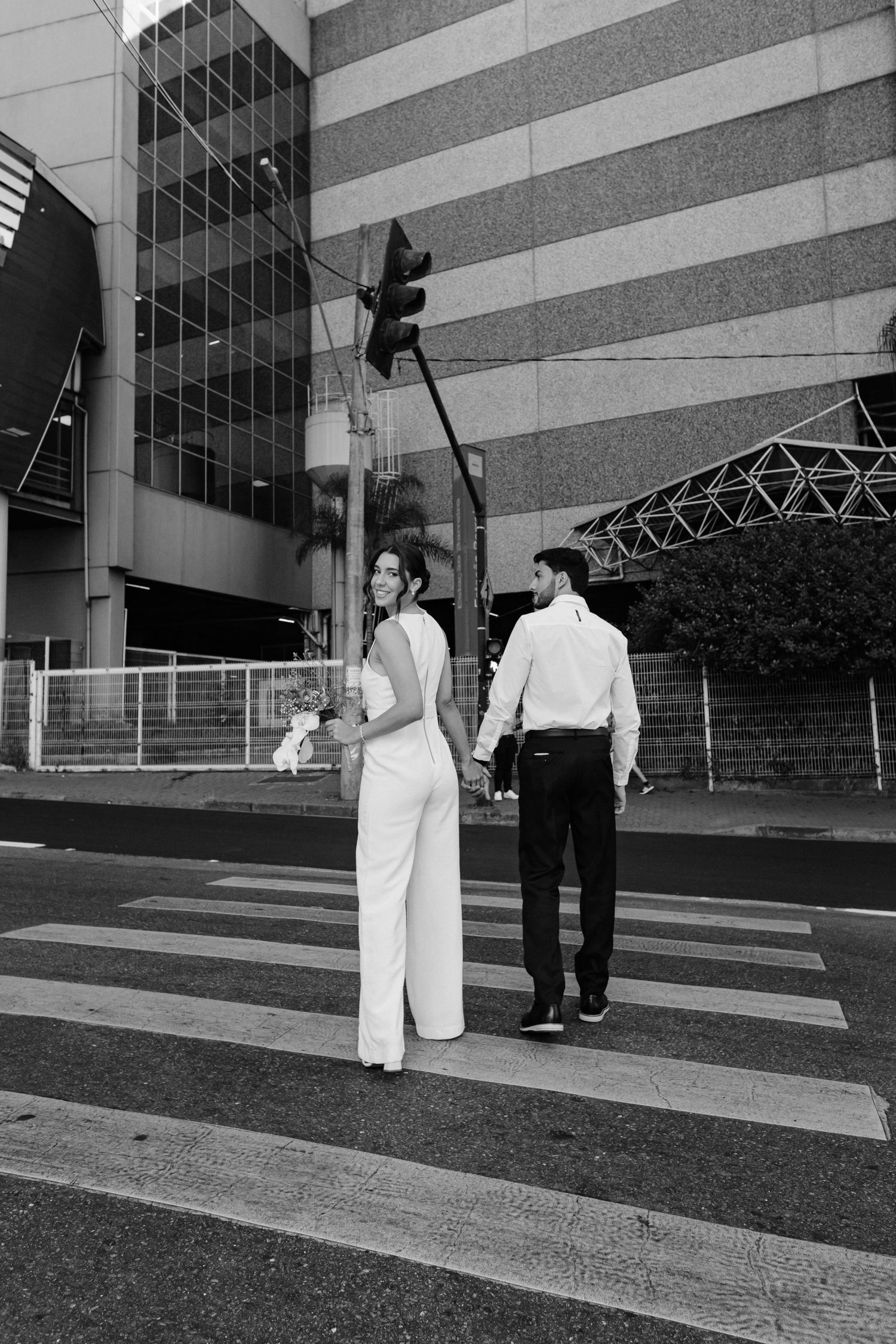Black and white photo of a couple holding hands while walking across a zebra crossing near a modern building.