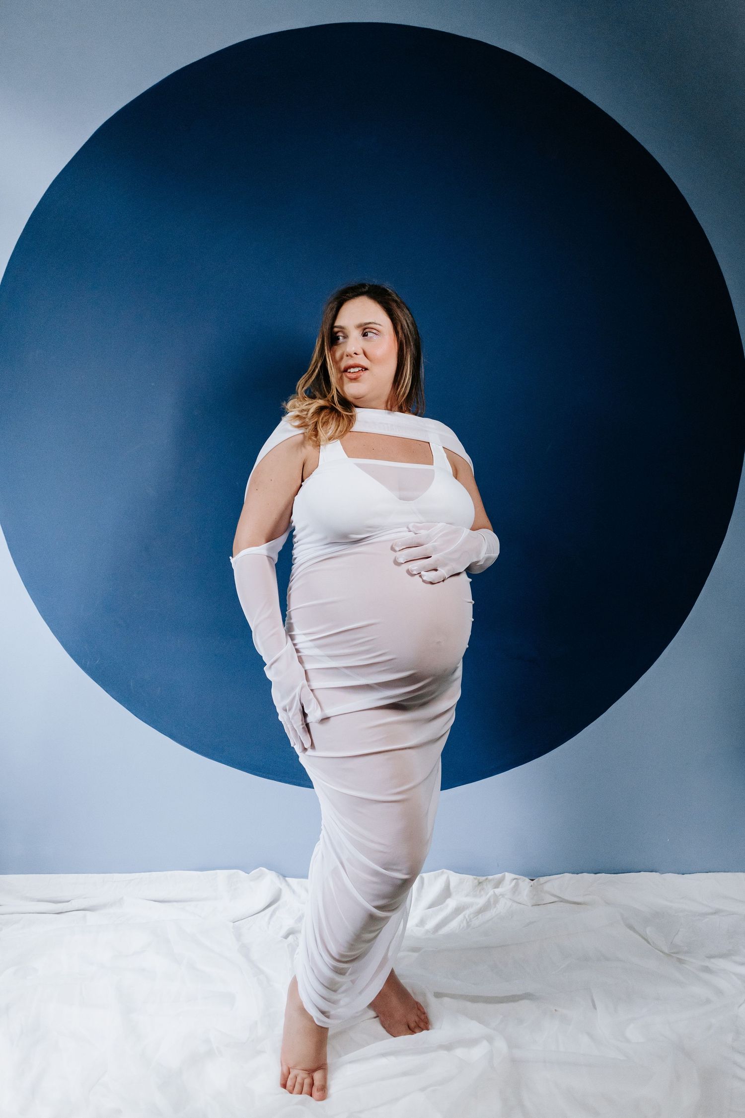 An expectant figure poses in a white gown against a bold blue circular backdrop.