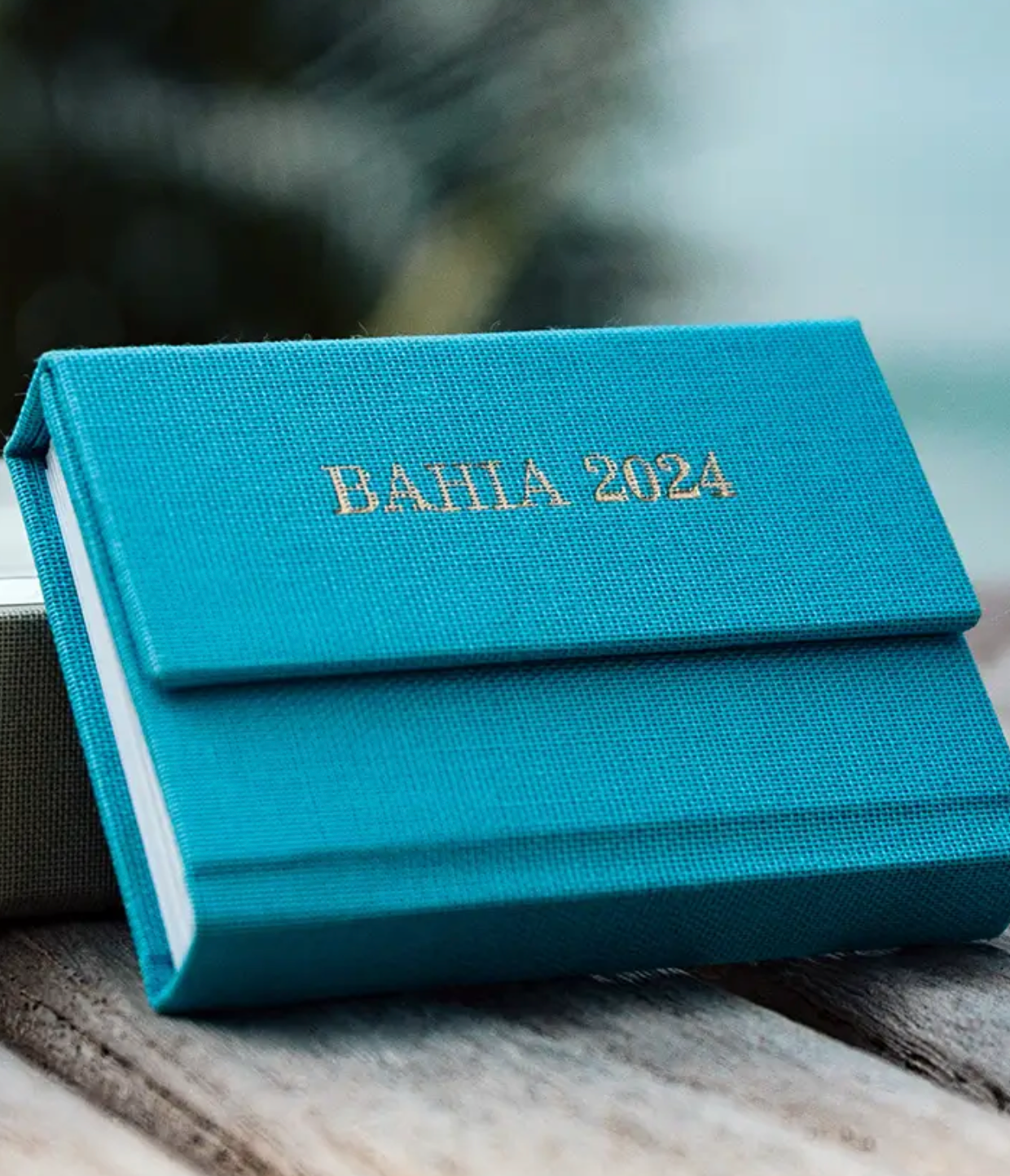 Teal linen-bound travel journal embossed with gold text reading BAHIA 2024, placed on a wooden surface.