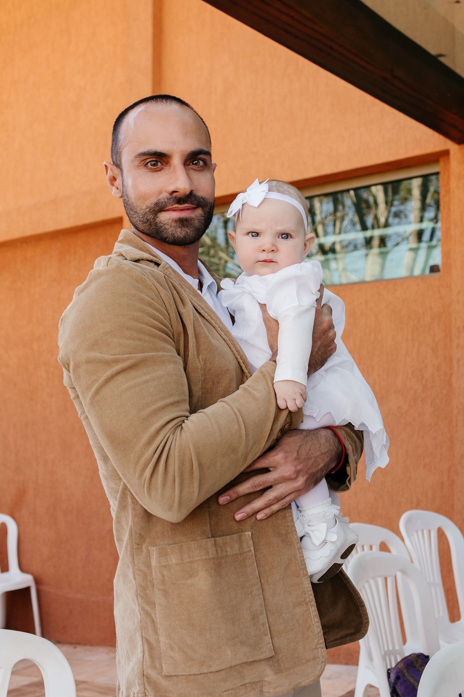 A man in a tan jacket holds a baby wearing white christening attire in three similar poses against an orange wall.