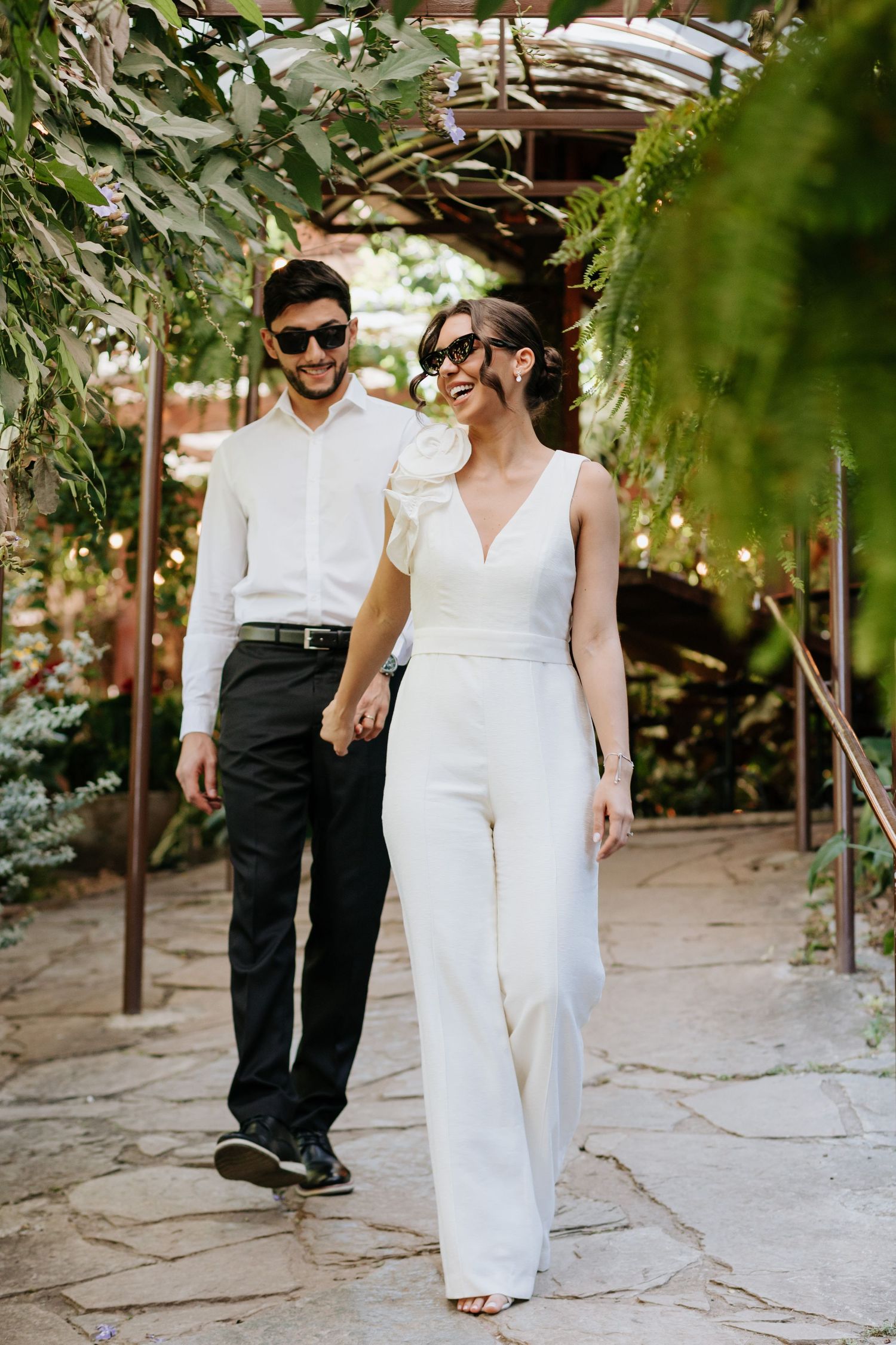 Couple in white jumpsuit and black pants holding hands walking through a lush garden pergola wearing sunglasses.