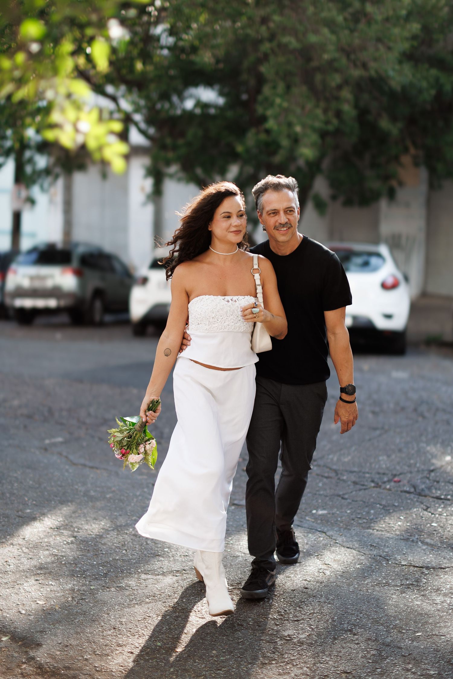 A smiling couple walks outdoors, woman in white outfit holding flowers, man in black shirt on a sunny street.