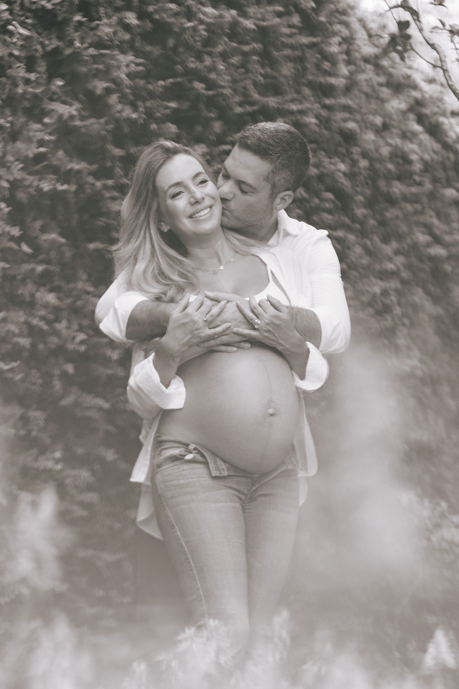 A romantic maternity photo in black and white shows an expecting couple embracing outdoors with natural foliage in background.