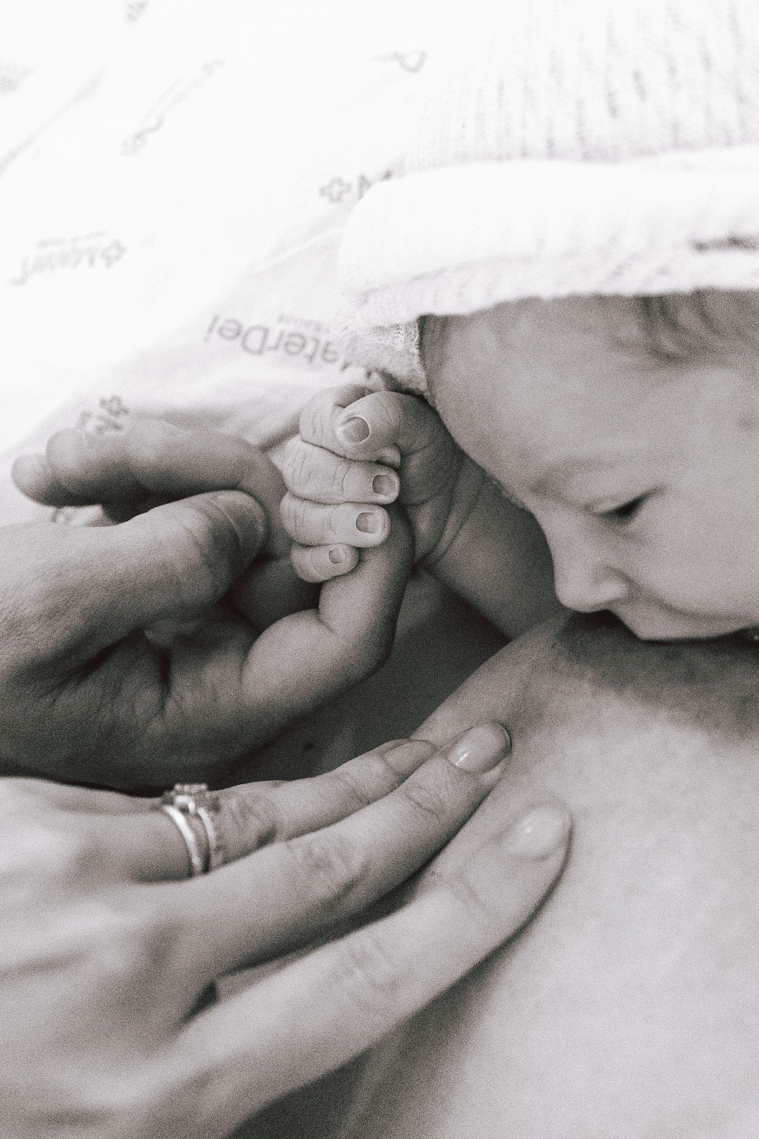 Black and white image of a newborn baby holding an adult's finger while wrapped in a white blanket.