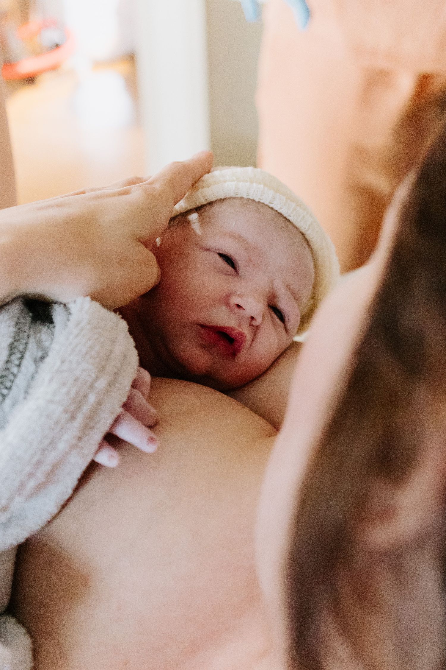 A newborn baby in a white knit hat being held against mother's chest during first breastfeeding moments after birth.