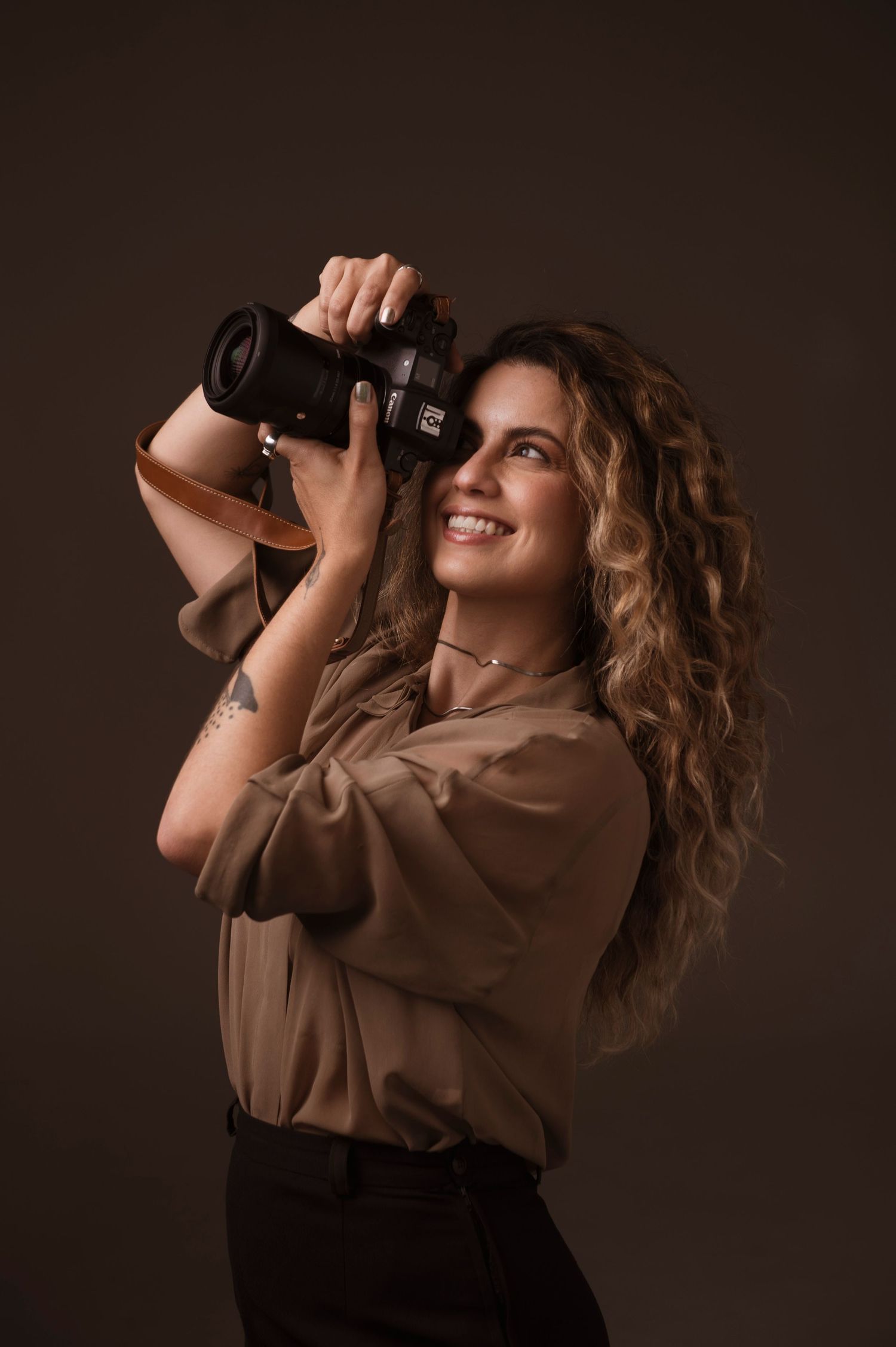 A photographer with wavy hair smiles while holding a DSLR camera against a dark brown background.
