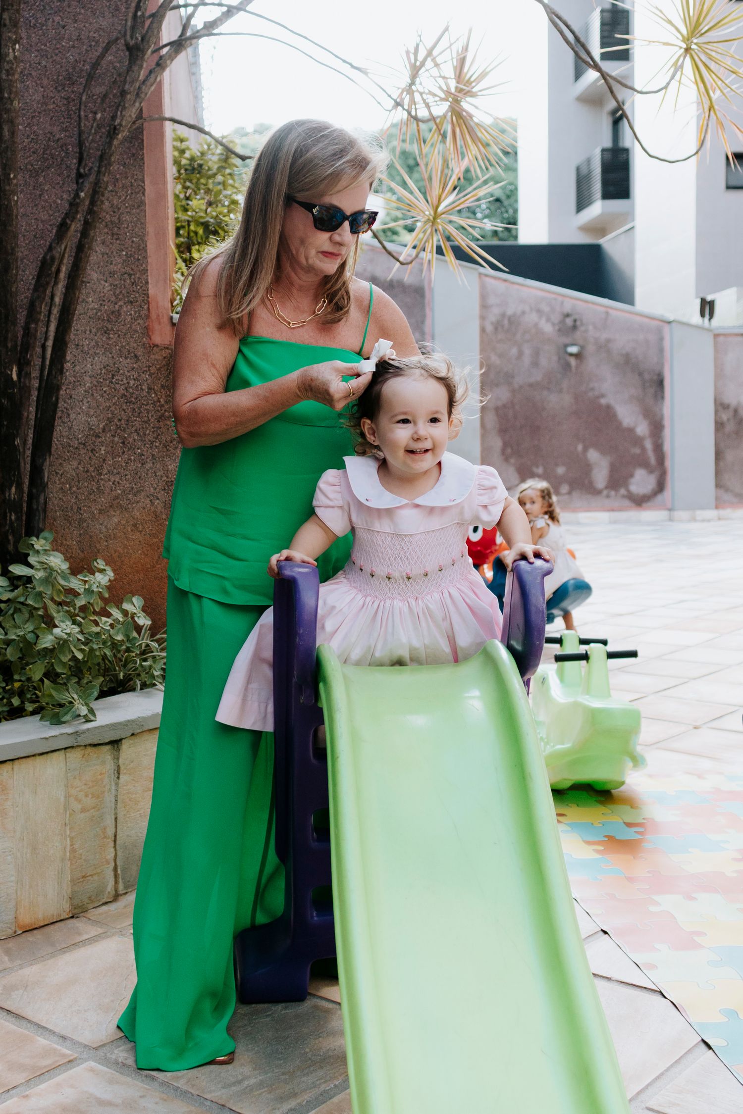 A woman in a green jumpsuit helps a child in a pink dress go down a slide.