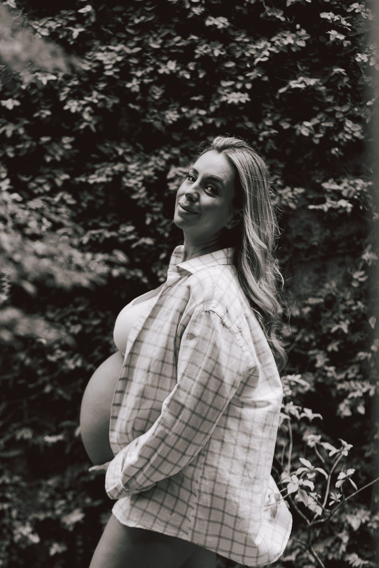 Black and white portrait of an expectant mother in a plaid shirt posing outdoors against leafy foliage.