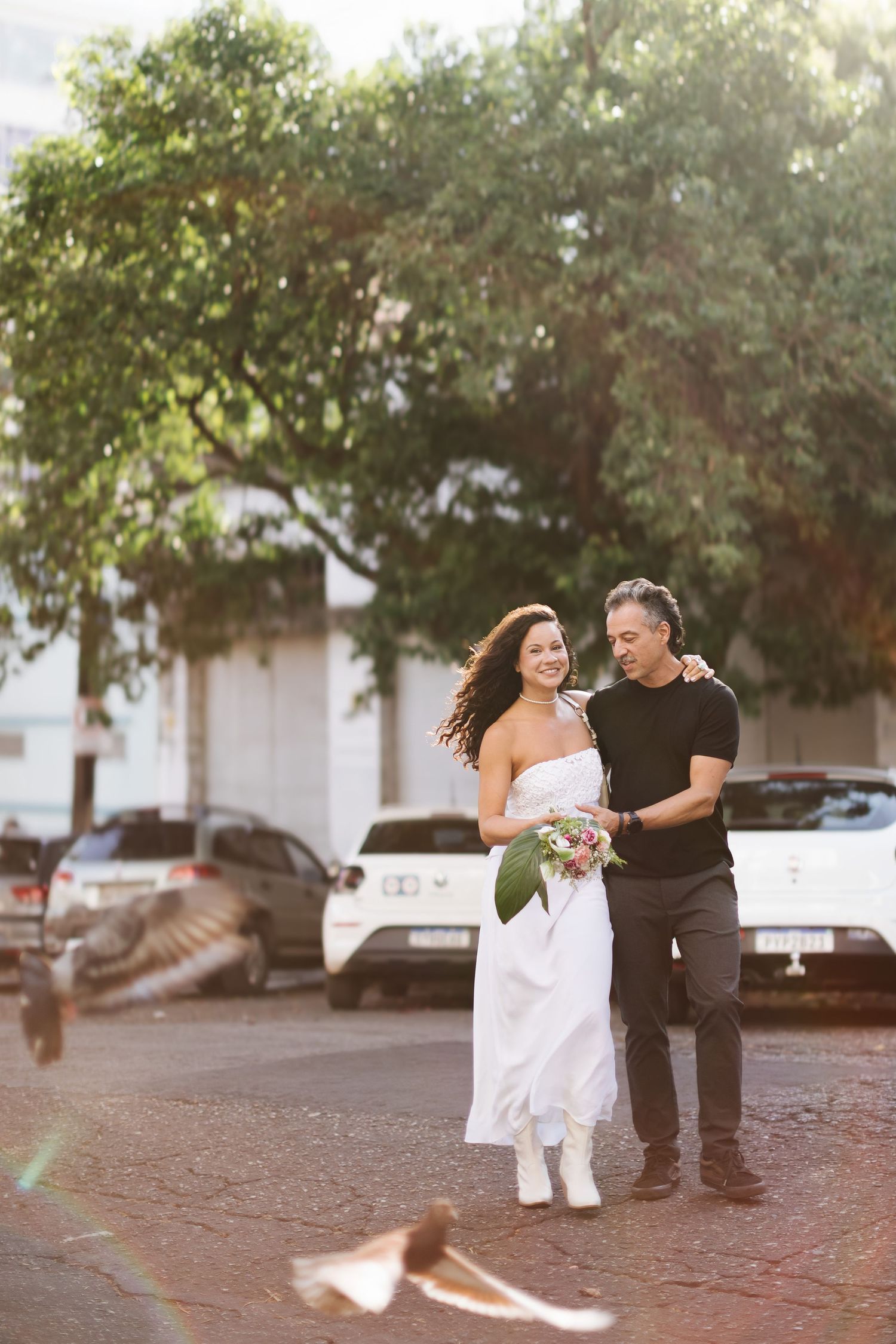 Bride in white dress holds bouquet as groom in black embraces her outdoors on a sunny cobblestone street.