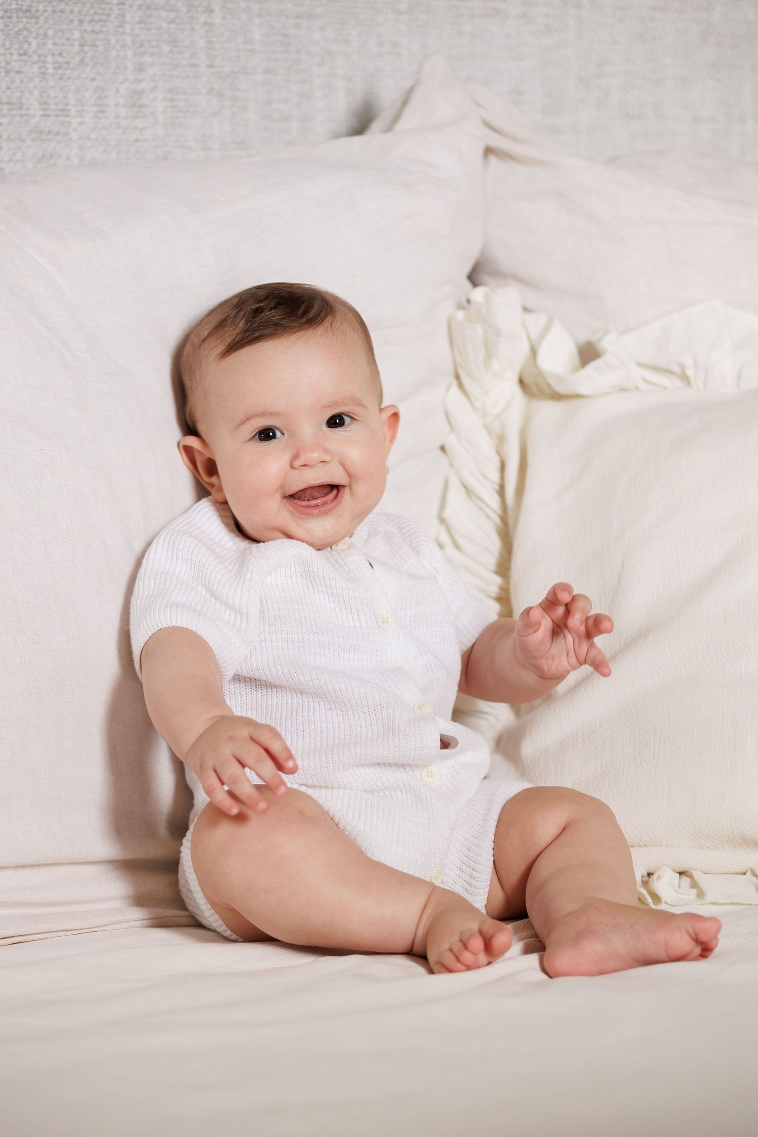 Smiling baby in white onesie sitting on a white bed with pillows, looking happy and reaching out.
